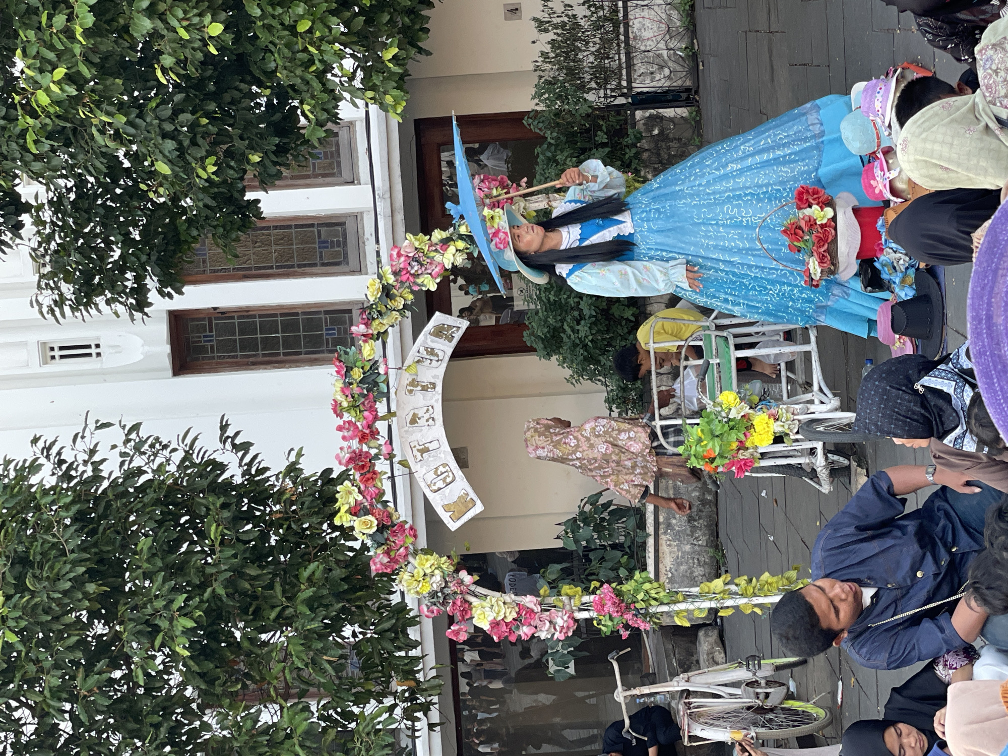 A woman dressed in a blue, ruffled, Victorian-style gown and a wide-brimmed hat stands holding a blue umbrella adorned with flowers, likely in the city of West Jakarta, Indonesia. She appears to be a performer or vendor, with a basket of red flowers at her feet, surrounded by a collection of hats on the ground. Behind her, an archway decorated with colorful artificial flowers frames a building with large windows. A young boy in a yellow shirt is seated in a white stroller, and another person in a floral hijab is walking away. Several other people are visible in the background, some of whom are seated on the ground. Bicycles are also present in the scene. The setting appears to be an outdoor public space, possibly a historical or cultural area, given the architecture and the presence of street performers or vendors.