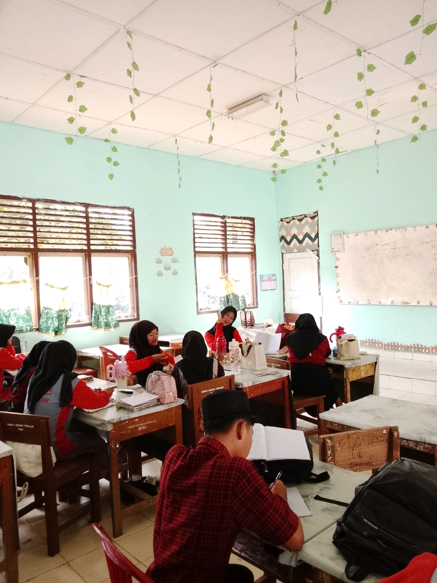 The image depicts an indoor scene, likely a classroom in Tanjung Setia, Indonesia. Several students, predominantly female wearing hijabs and red and gray uniforms, are seated at wooden desks, engaged in various activities. Some are writing or reading, while others appear to be interacting with one another. A male student in a red checkered shirt and black peci hat is also visible, writing in a notebook.
The classroom walls are painted a light green, and the ceiling is a white tiled suspended grid, decorated with artificial green ivy garlands. Windows with wooden blinds are present on one wall, with green curtains visible. A whiteboard with some writing is mounted on another wall. Other objects in the scene include books, notebooks, water bottles, a pink cup, and backpacks. The overall impression is a typical classroom environment during a lesson or study session.