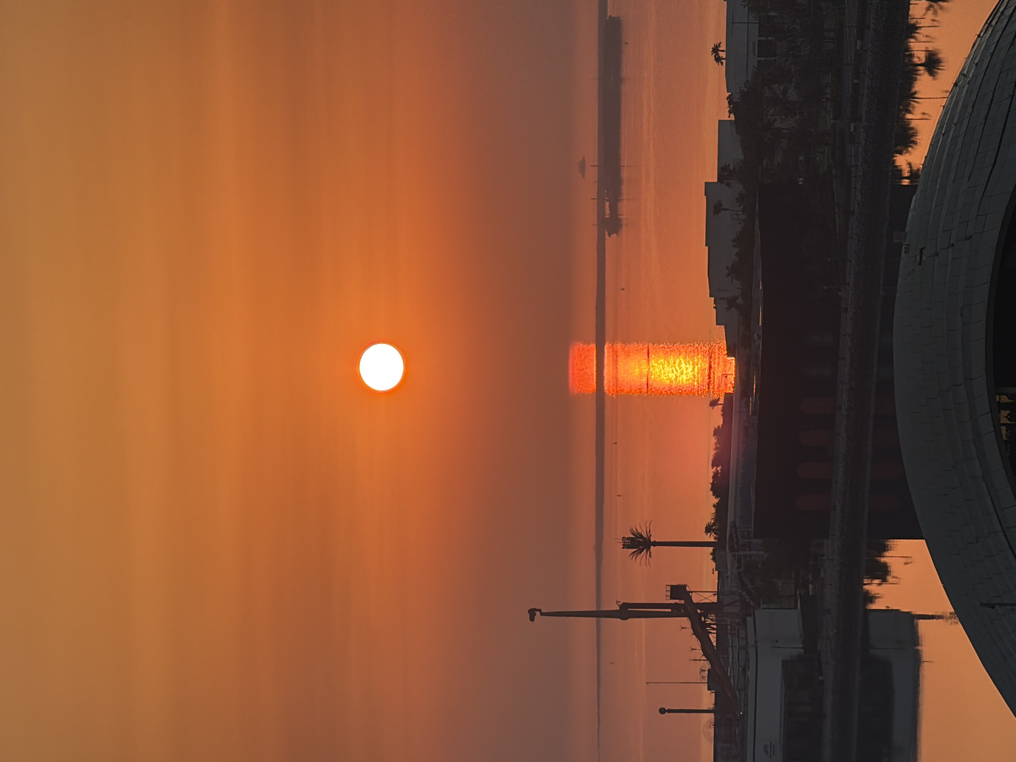 The image depicts a coastal urban scene at sunrise or sunset. A large, bright orange sun hangs low in the sky, casting a warm glow and a shimmering reflection on the water. The sky transitions from a bright orange near the sun to a softer, lighter orange towards the top of the frame.
In the middle ground, a body of water separates the viewer from the cityscape. Several vessels are visible on the water, including a large ship anchored in the distance and what appears to be a barge or cargo ship closer to the shore. Structures along the waterfront include industrial-looking buildings with dark facades, some with arched entrances. A tall, slender tower with a light fixture is prominent on the left, alongside what looks like a water slide or amusement park ride. A palm tree, silhouetted against the sky, stands to the right of the tower.
In the foreground, the top of a large, curved, gray structure is visible, suggesting a building with a distinctive architectural feature. Reflections of palm trees and buildings are visible in the calm water. The overall impression is one of a harbor or industrial waterfront bathed in the dramatic light of a sunrise or sunset. The location is identified as Unknown, Unknown.