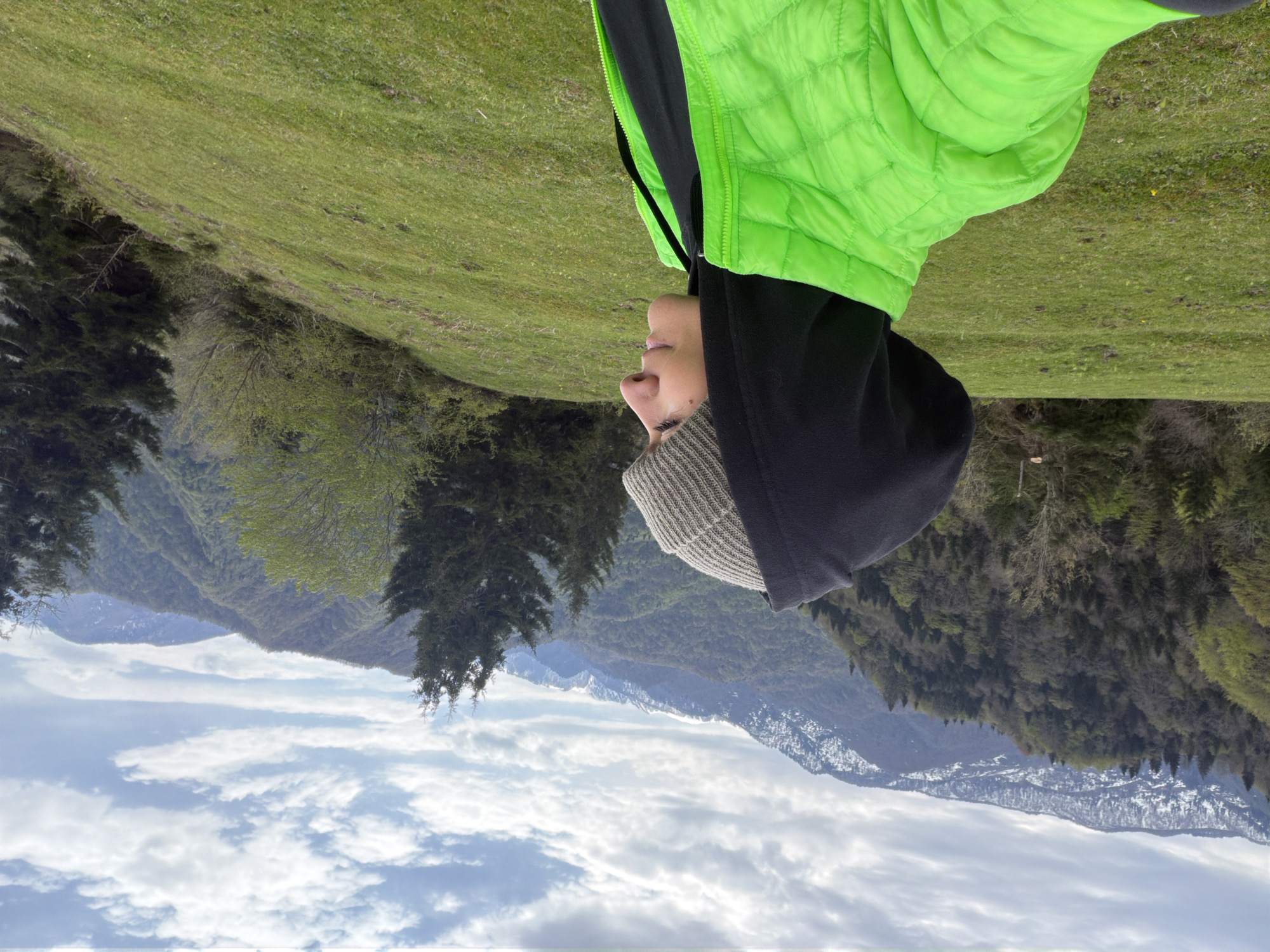 A person wearing a gray beanie, black hooded sweatshirt, and a bright green puffer vest is centered in the image, framed by a landscape. The background consists of rolling green hills, some covered in trees, with snow-capped mountains visible in the distance beneath a partly cloudy sky. The exact location is not identifiable from the image.