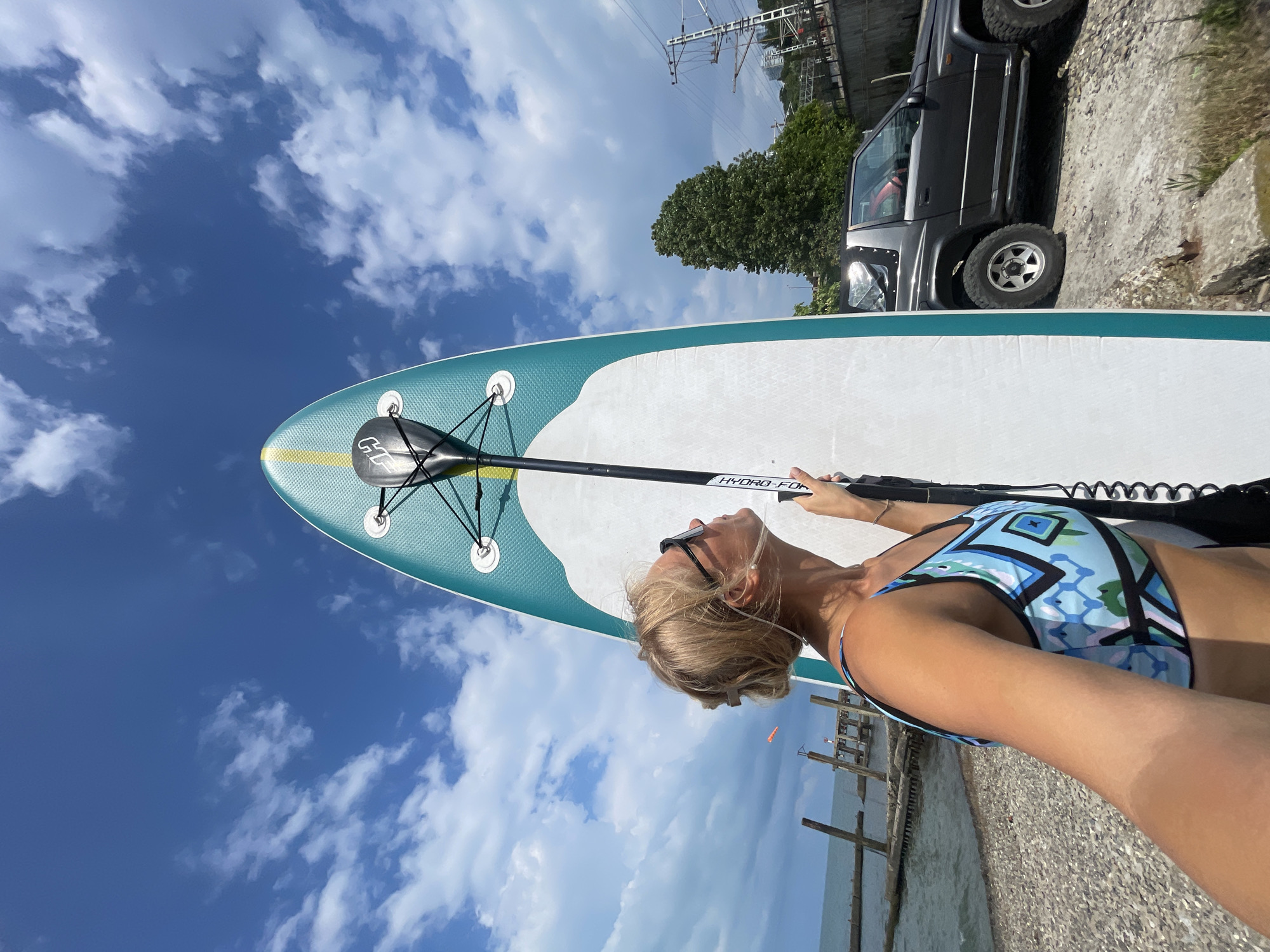 A woman stands on a beach, holding a paddleboard and paddle, looking up at the sky. She is wearing sunglasses and a blue and white patterned bikini. The paddleboard is white with a turquoise and yellow design along the edge. In the background, a dark grey SUV is parked on a gravelly surface, and a line of trees and power lines can be seen. The scene is set against a bright blue sky with scattered white clouds. The sun is shining, suggesting it is daytime. The water and a wooden pier are visible in the lower right of the frame, indicating a coastal or lakeside location. The woman's gaze is directed upwards, perhaps enjoying the weather or anticipating an activity.