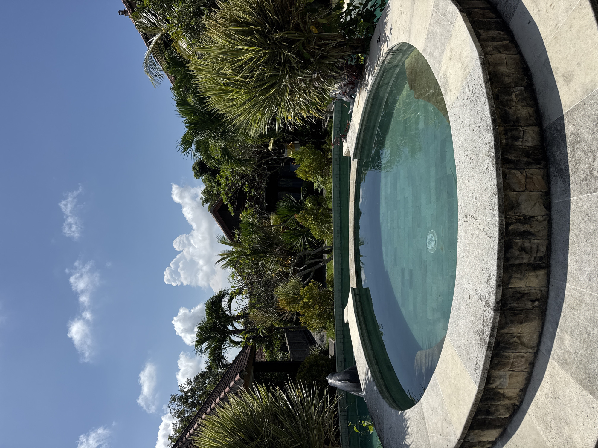 A round pool with tiled green interior is bordered by grey stone. Stairs are inset in the stone around the pool. Tropical vegetation surrounds the pool, which is in a garden setting. A blue sky with scattered clouds is visible above the vegetation. The location is Kuta Selatan, Indonesia.