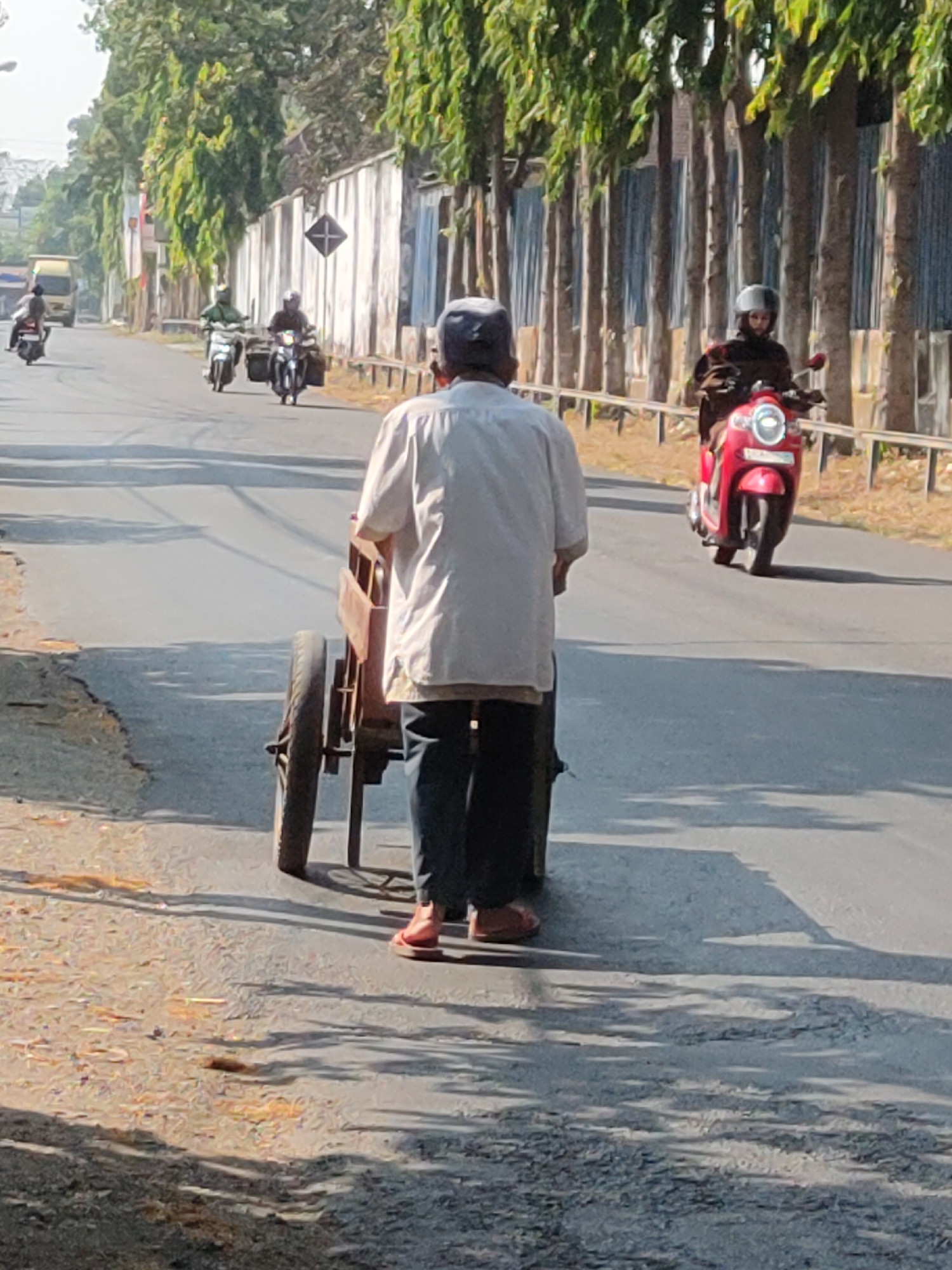 The image shows a street scene in Kediri City, Indonesia. In the foreground, a person wearing a white shirt and dark pants pushes a wooden cart with two wheels. The cart appears to be made of aged wood and is positioned behind the person. The person is facing away from the camera. The street is paved, and there is a rougher dirt and gravel area to the left.
In the background, several motorcycles are visible on the road. One motorcycle is red and is being ridden by a person wearing a helmet. Another motorcycle is dark and is being ridden by a person wearing a helmet and a green shirt. Further down the road, another motorcycle is visible, and a yellow truck is in the distance on the left side of the road.
A row of trees with green foliage lines the right side of the road, with a metal fence or barrier in front of them. Behind the trees, a white wall with vertical elements can be seen. The sky is bright and appears to be daytime. The overall impression is that of a busy street in an urban or semi-urban area in Indonesia.