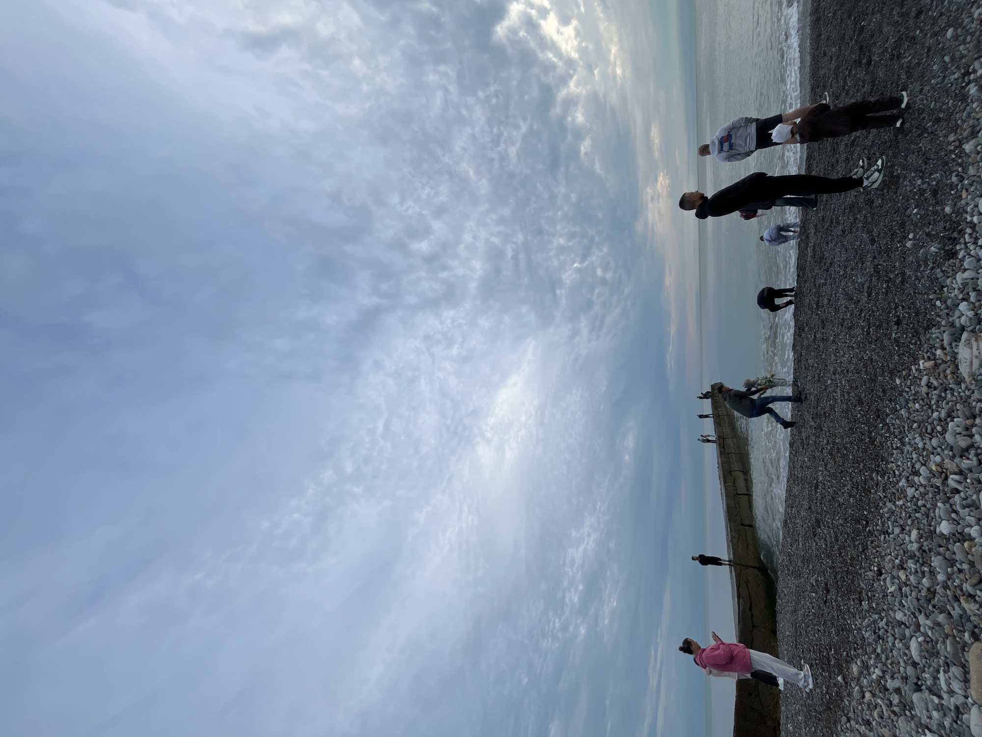 The image, taken in Sochi, Russia, depicts a tranquil coastal scene under an overcast sky. Multiple individuals are scattered across a pebble beach and along concrete structures adjacent to the sea. The lighting is diffused, suggesting either early morning, late afternoon, or a generally cloudy day.
In the foreground, a person dressed in a pink hoodie and white pants stands on a concrete barrier or pier, facing the water. Further along this structure, another individual in dark attire is visible. On the dark pebble beach, several other people are present. A group closer to the foreground includes one person in a grey and white hoodie standing alongside another in a dark jacket and white cap. Other figures are seen nearer the water's edge; some are bending over, while others stand upright, seemingly observing the calm sea. In the mid-ground, a person in a grey shirt and jeans is standing on a concrete section of the shore, close to the water.
The overall activity appears leisurely, with people enjoying a casual stroll or simply taking in the view of the sea. The setting features a distinct pebble beach and man-made concrete elements extending into or alongside the water, characteristic of coastal areas. There is no visible text in the image.