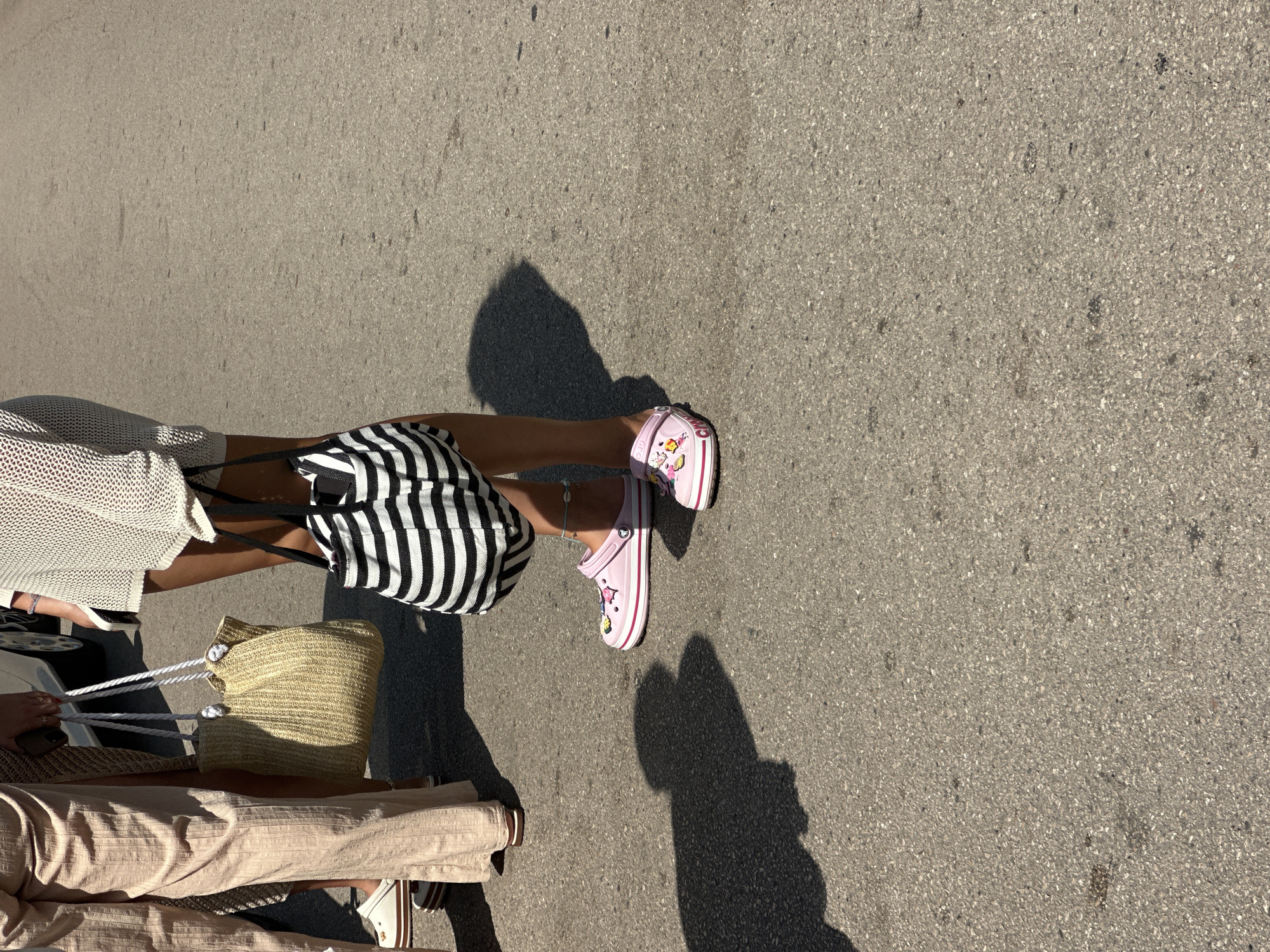 The image shows a close-up view of the lower bodies of three individuals standing on an asphalt surface, possibly a street or sidewalk, in Bečići, Montenegro. The lighting suggests it is daytime, with distinct shadows cast on the ground.
The person on the left is wearing loose-fitting, light-colored pants that extend to their ankles, and white platform sandals with a brown sole. They are carrying a light-brown woven straw tote bag with white rope handles.
The person in the middle is wearing a white, textured knitted dress or cover-up that falls to their mid-thigh. They are carrying a black and white striped tote bag. On their feet, they are wearing pink platform Crocs adorned with colorful charms. A delicate anklet is visible on their left ankle.
The third person, partially visible on the right, is also wearing pink platform Crocs with charms.
The background is out of focus but suggests a sunny outdoor environment, possibly near a building or parked vehicle, given the presence of a shadow that appears to be cast by a car. The asphalt surface is grey and shows some small dark debris.