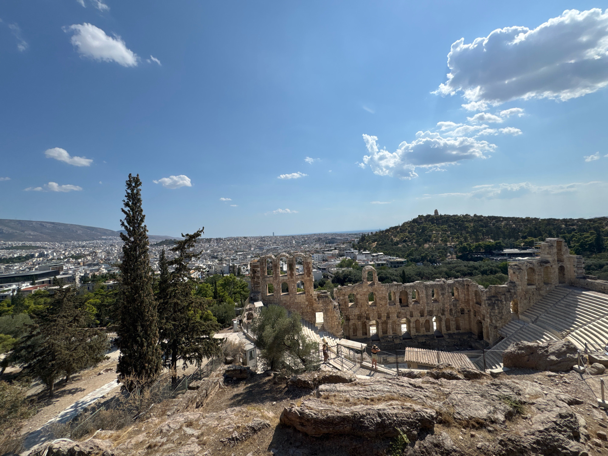 The image captures a wide-angle view of the Odeon of Herodes Atticus in Athens, Greece, on a sunny day. In the foreground, rocky terrain with sparse vegetation and a tall, slender evergreen tree on the left dominate. Beyond this, the ancient stone structure of the Odeon is prominent, with its multi-tiered seating and remnants of its stage building. Two people are visible near the entrance to the seating area.
The middle ground features more of the Odeon's ruins and pathways, interspersed with green trees and fencing. A dense urban landscape, characteristic of Athens, stretches into the background under a clear blue sky dotted with white clouds. The city extends towards a distant, hazy mountain range. The overall scene depicts a historical landmark within a bustling modern city.