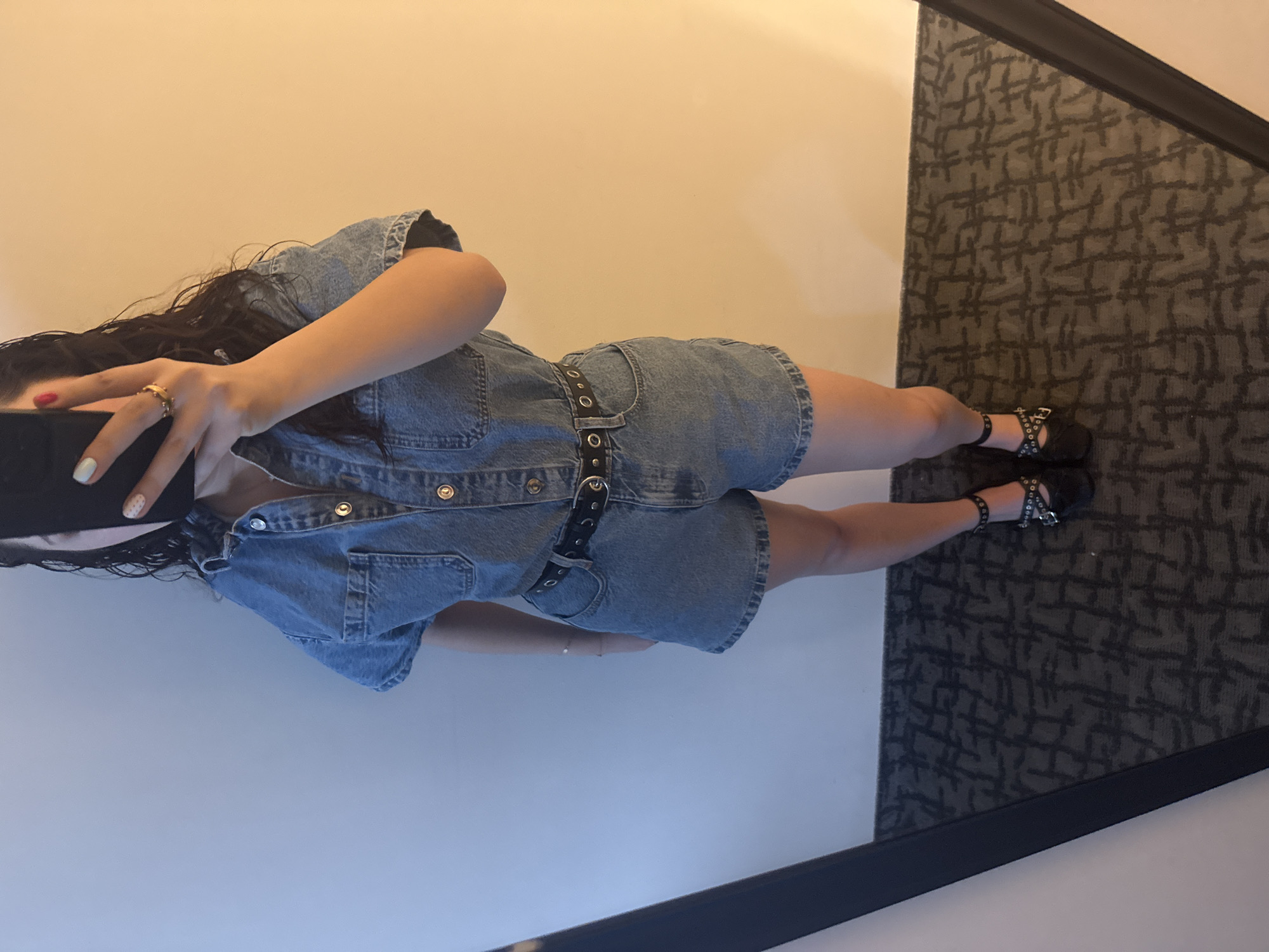 A person wearing a denim romper, a black belt, and black shoes with ankle straps is standing on a dark patterned carpet against a light beige wall. The person is holding a black object, possibly a phone, up to their face, obscuring it. The photo appears to be taken in a mirror, as indicated by the frame around the carpet. The location is Mexico City, Mexico, based on information not visible in the image.