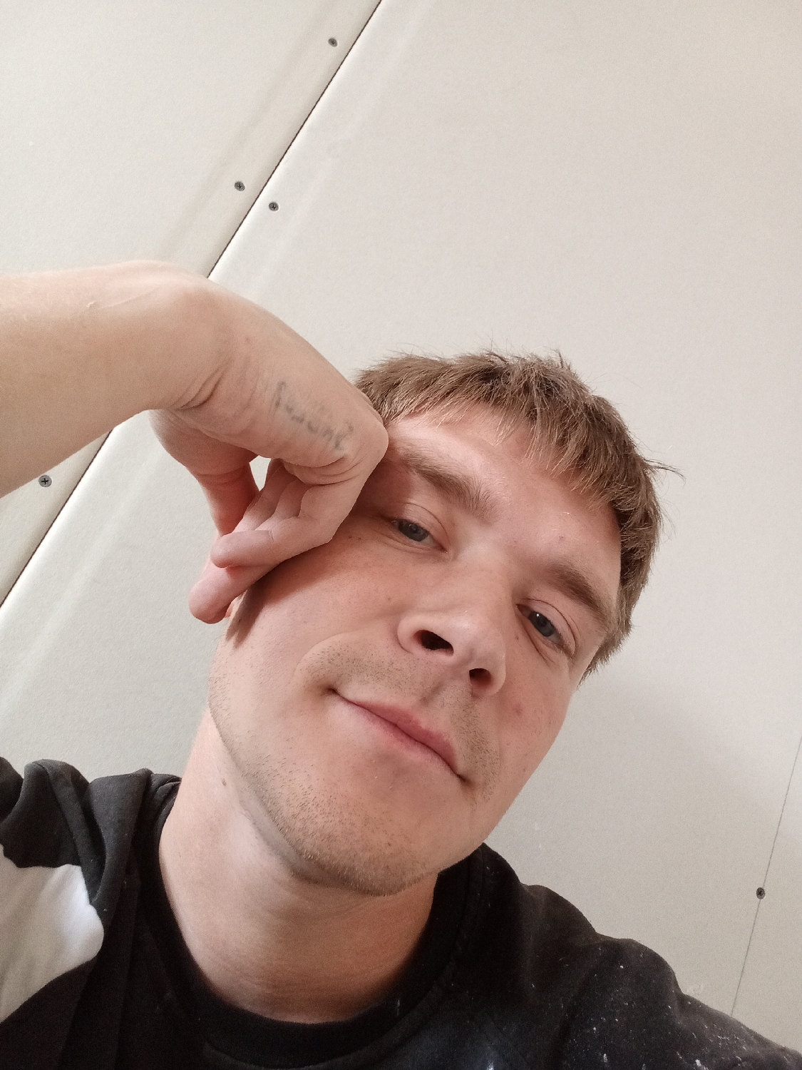 A young Caucasian man is captured in a close-up, low-angle selfie. He has light brown, slightly disheveled hair and light-colored eyes. A short stubble covers his chin and jawline. He is wearing a dark, long-sleeved top, possibly a sweatshirt, which features a white pattern on the left side and appears to be speckled with light-colored debris, resembling paint.
His left hand is raised, with his fingers resting on his forehead and his thumb near his temple, conveying a casual or thoughtful pose. On the back of his left hand, near the knuckles, a faded and largely illegible text tattoo is visible. The man looks directly at the camera with a neutral to slightly content expression, featuring a subtle, closed-mouth smile.
The background consists of plain, light-colored panels, likely drywall or similar interior construction, with visible seams and screws. This suggests an indoor setting, possibly a room under construction or a utilitarian space. No specific time of day or external location cues are discernible from the image.
