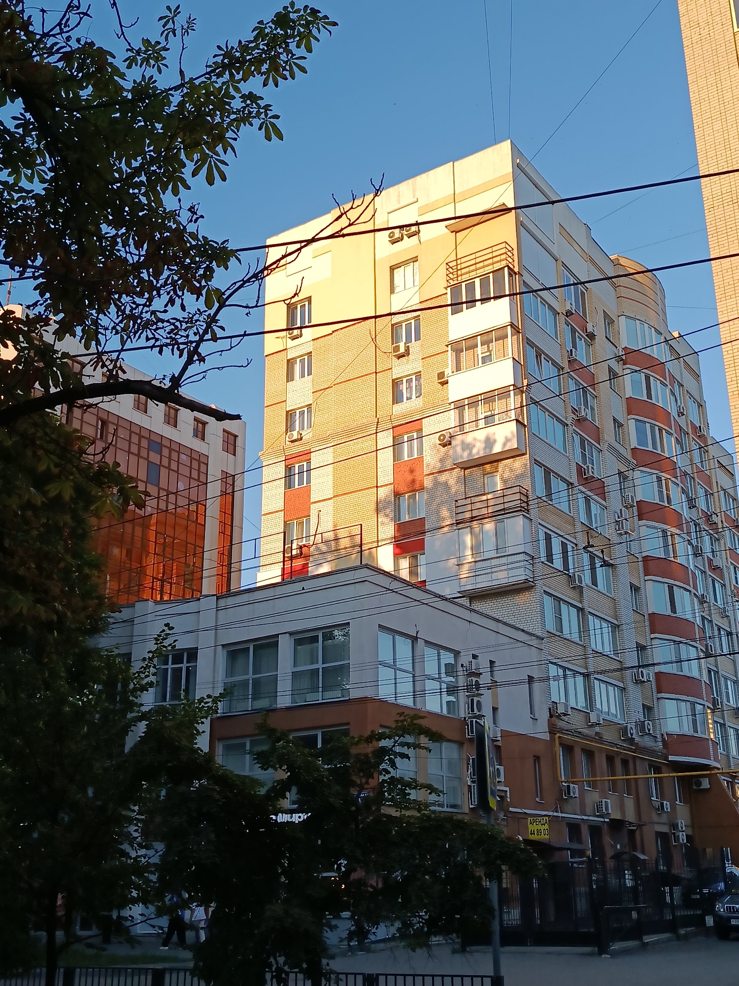 The image captures a modern residential building in Saratov, Russia, bathed in the warm light of the late afternoon sun. The building, characterized by its light yellow brick facade and curved red and white sections, features numerous windows and balconies, many equipped with air conditioning units. In front of this main structure, a slightly lower building with a white facade and large windows is visible, reflecting the sunlight. To the left, another building with a glass facade, tinted in orange, stands tall. Overhead, a network of electrical wires crisscrosses the sky, partially obscuring the buildings. The lower part of the image shows trees with green foliage, a black metal fence, and a street with a few parked cars. The sky is a clear, bright blue.