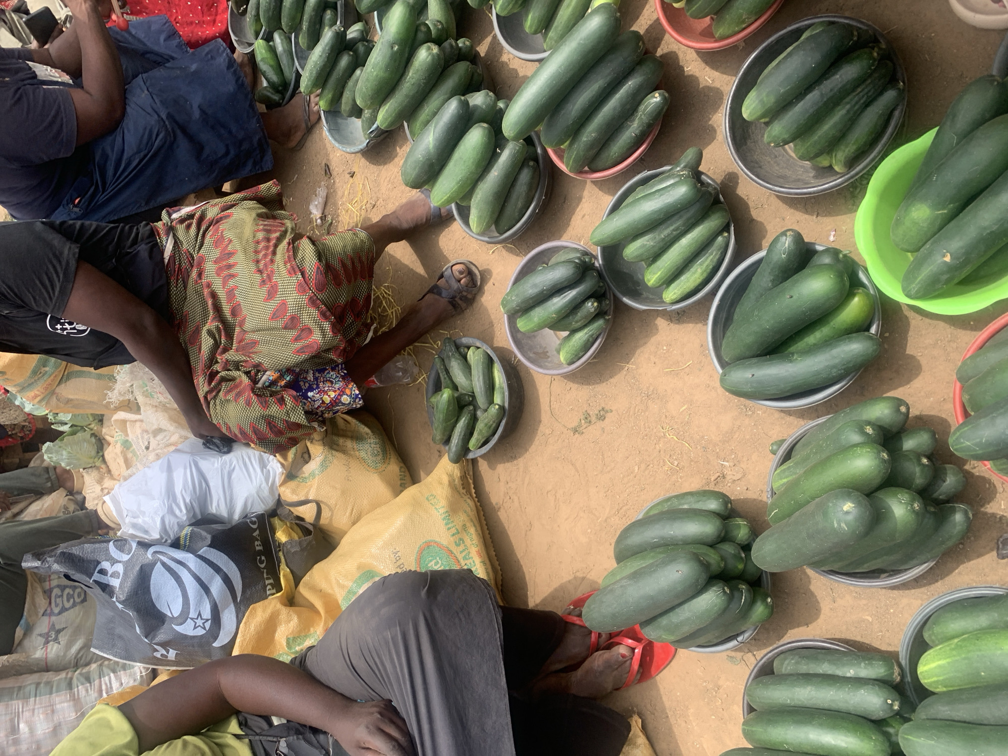 The image depicts a marketplace scene in Jos, Nigeria. Several individuals are seated on bags and the ground among numerous bowls filled with cucumbers. The ground is a light-brown dirt surface. People wear casual clothing, and the bowls are various colors (grey, green, red). The scene has a ground-level perspective with natural daylight.