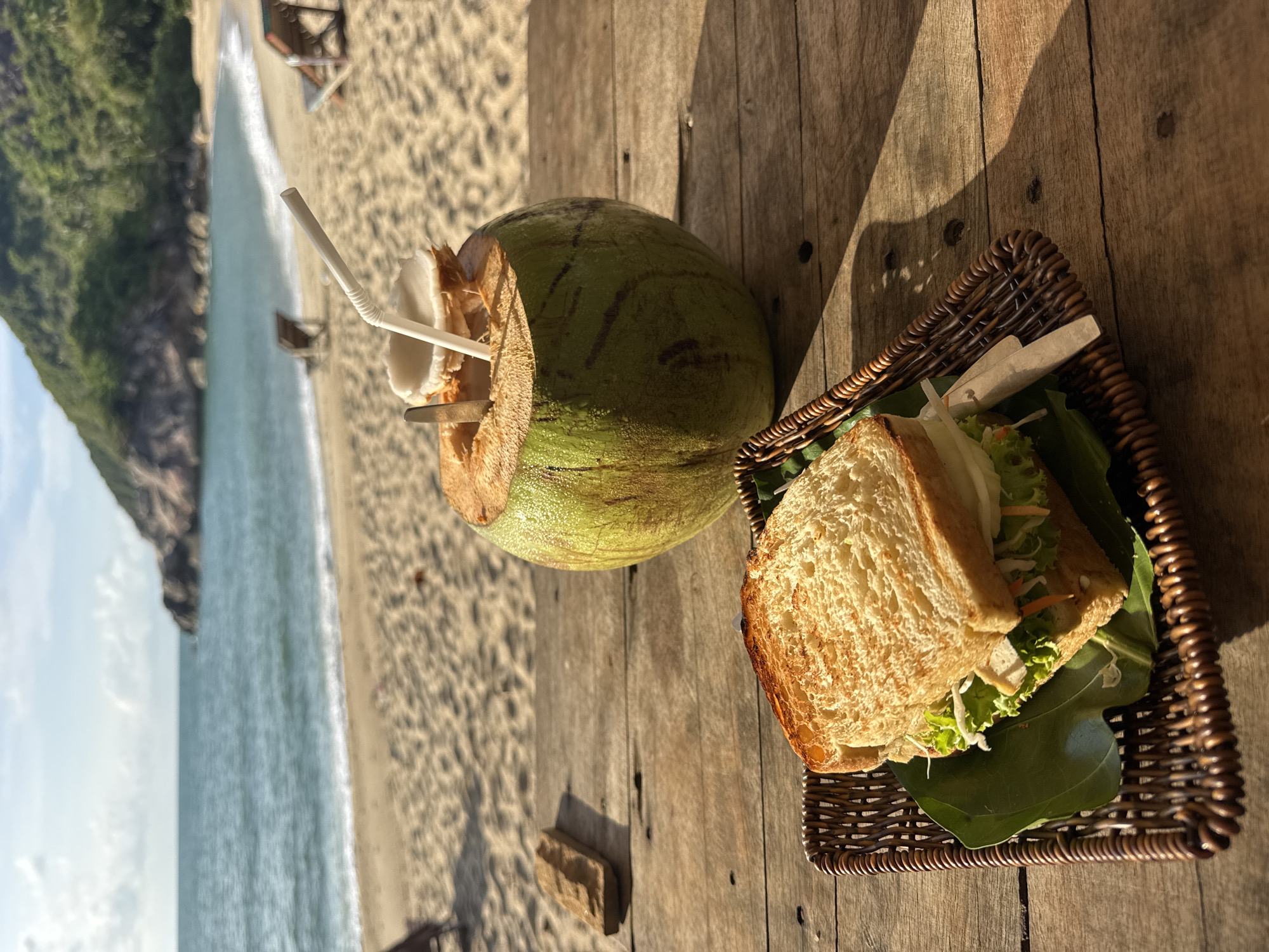 On a wooden table, a scene unfolds with a coconut beverage and a sandwich in a woven basket. The coconut, topped with a white straw, sits next to the basket containing a sandwich filled with lettuce and other ingredients. In the background, a beach and ocean view are visible, with a mountainous coastal landscape. The location is Baan Thong Nang, Thailand.