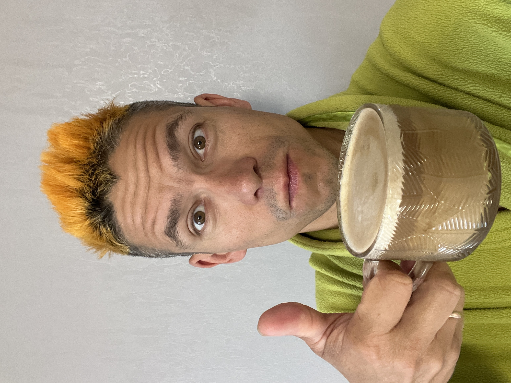 A man with spiky orange hair is holding a clear glass mug filled with a frothy beverage and giving a thumbs-up. He is wearing a bright green bathrobe. The background is a plain, textured light gray wall. The man's expression is one of surprise, with wide eyes and a slightly open mouth. The image appears to be a selfie taken indoors. There is no visible indication of the city of Chaikovsky, Russia, in the image.