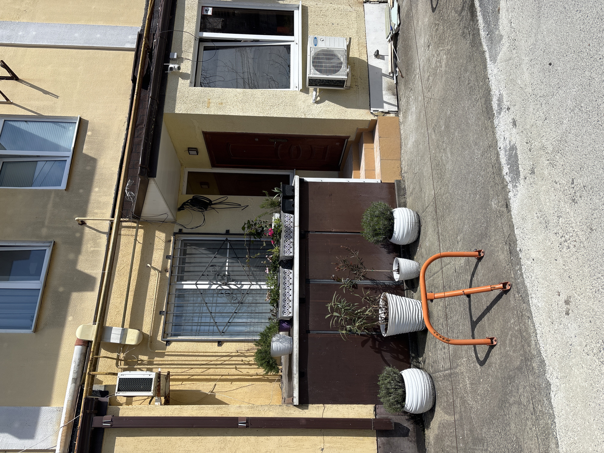 The image depicts the facade of a building in Sochi, Russia, as suggested by architectural style and climate. The building is two-story and painted yellow. There are windows on both floors, with a security grille on the lower-level window. The front door is dark wood. Several air conditioning units are visible on the exterior. The facade is lined with potted plants and a rack is present. The area in front of the building appears to be a pedestrian zone.