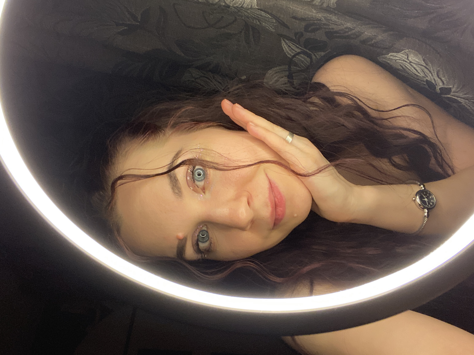 A woman with long, wavy brown hair is posing in what appears to be an indoor setting. Her eyes are light blue and she has glitter makeup around them. She's wearing a silver ring on her finger and a small, round watch on her wrist. Her hand is raised near her face. She is framed by a bright ring light, and a dark patterned fabric is visible in the background. The location is stated as being in the city of Chaikovsky, Russia.