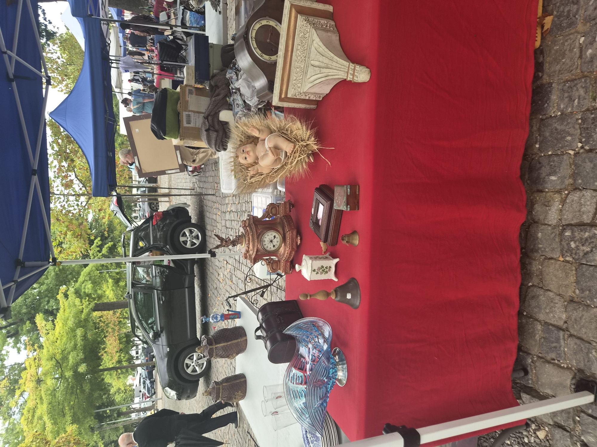 This image depicts an outdoor flea market in Barcelos, Portugal. A red tablecloth is spread over a table, displaying various items for sale. Among the items are decorative baskets, glassware, a wooden chest, a ceramic pot with a floral design, a bell, a doll resembling a baby in a straw crib, a decorative clock with an eagle finial, and a wall-mounted decorative bracket. In the background, a dark SUV is parked with its doors open, and several people are browsing at other stalls under blue tents. The ground is paved with cobblestones, and trees with green and yellow foliage line the background, suggesting an autumnal setting.