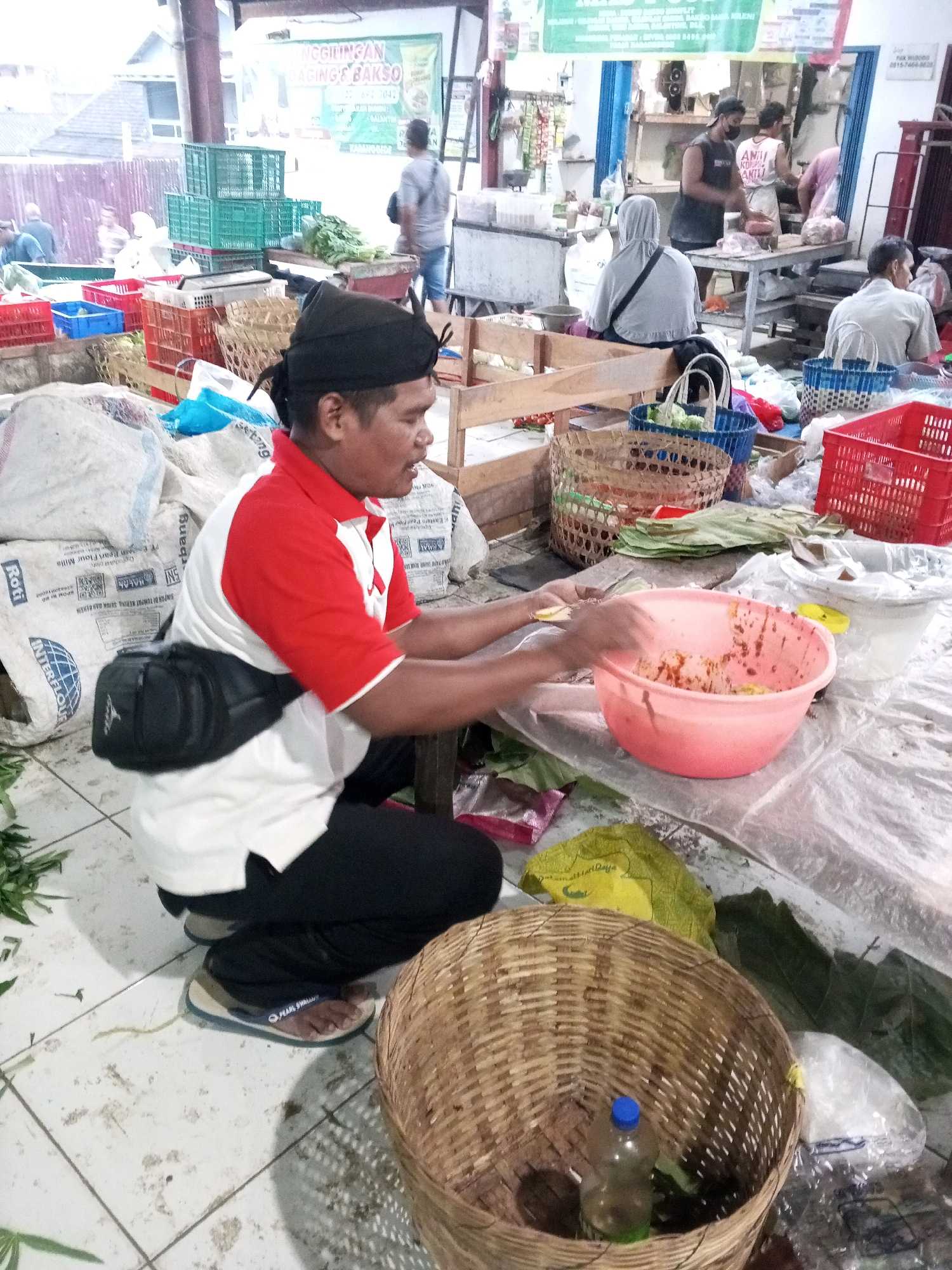The image depicts an outdoor market scene in Gentan, Indonesia. In the foreground, a man wearing a red and white shirt, black pants, and a black fanny pack is crouched down, actively mixing food in a large pink bowl with his hands. He has a black head covering. To his right, a woven basket sits on the ground, containing a plastic bottle with a blue cap and some green leaves. Behind him, large white sacks are stacked. In the background, various market stalls are visible, with displays of green crates, red plastic crates, and produce. Other individuals can be seen in the background, engaged in market activities. The ground is tiled and appears somewhat dirty.