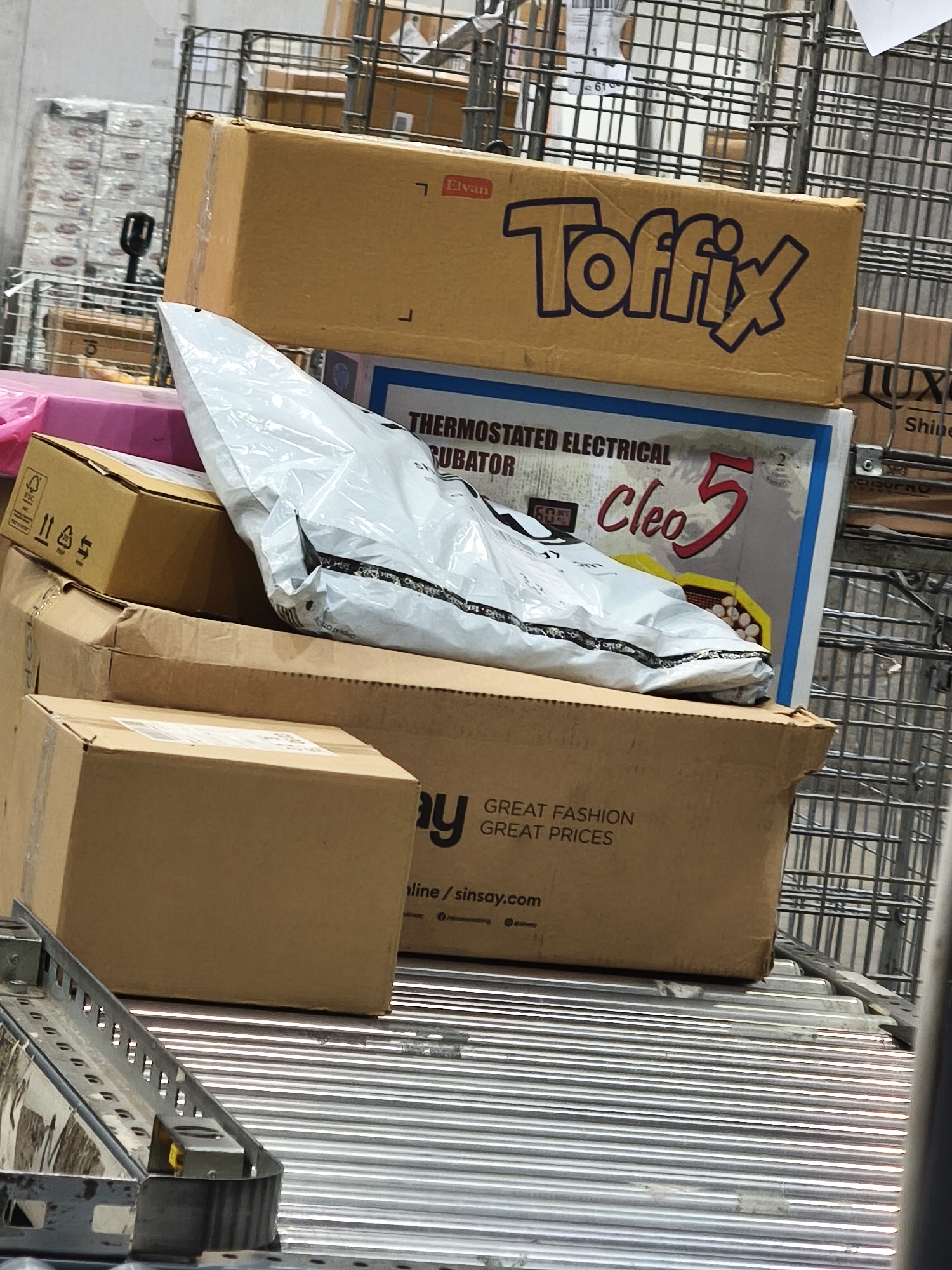 The image depicts a stack of packages and boxes in what appears to be a distribution center or warehouse setting. The primary objects include:
1. **Boxes:** Several cardboard boxes are stacked haphazardly. One prominently displays the brand "Toffix". Another is labeled "THERMOSTATED ELECTRICAL RUBATOR Cleo 5". A box with "GREAT FASHION GREAT PRICES line/sinsay.com" is also visible.
2. **Shipping Envelope:** A white, opaque shipping envelope lies on top of the boxes.
3. **Metal Rack:** A wire metal rack serves as a backdrop, suggesting the boxes are in a storage or transportation area.
4. **Conveyor belt:** A portion of a metal conveyor belt is visible in the foreground, indicating that the packages are likely in transit within a facility.
5. **Background details:** In the background, there are more indistinct packages wrapped in plastic, further reinforcing the impression of a distribution or storage facility.
There are no people visible in the image. The overall environment suggests a functional, industrial space. It is not possible to determine the precise location in Burgas, Bulgaria, from the given image alone.