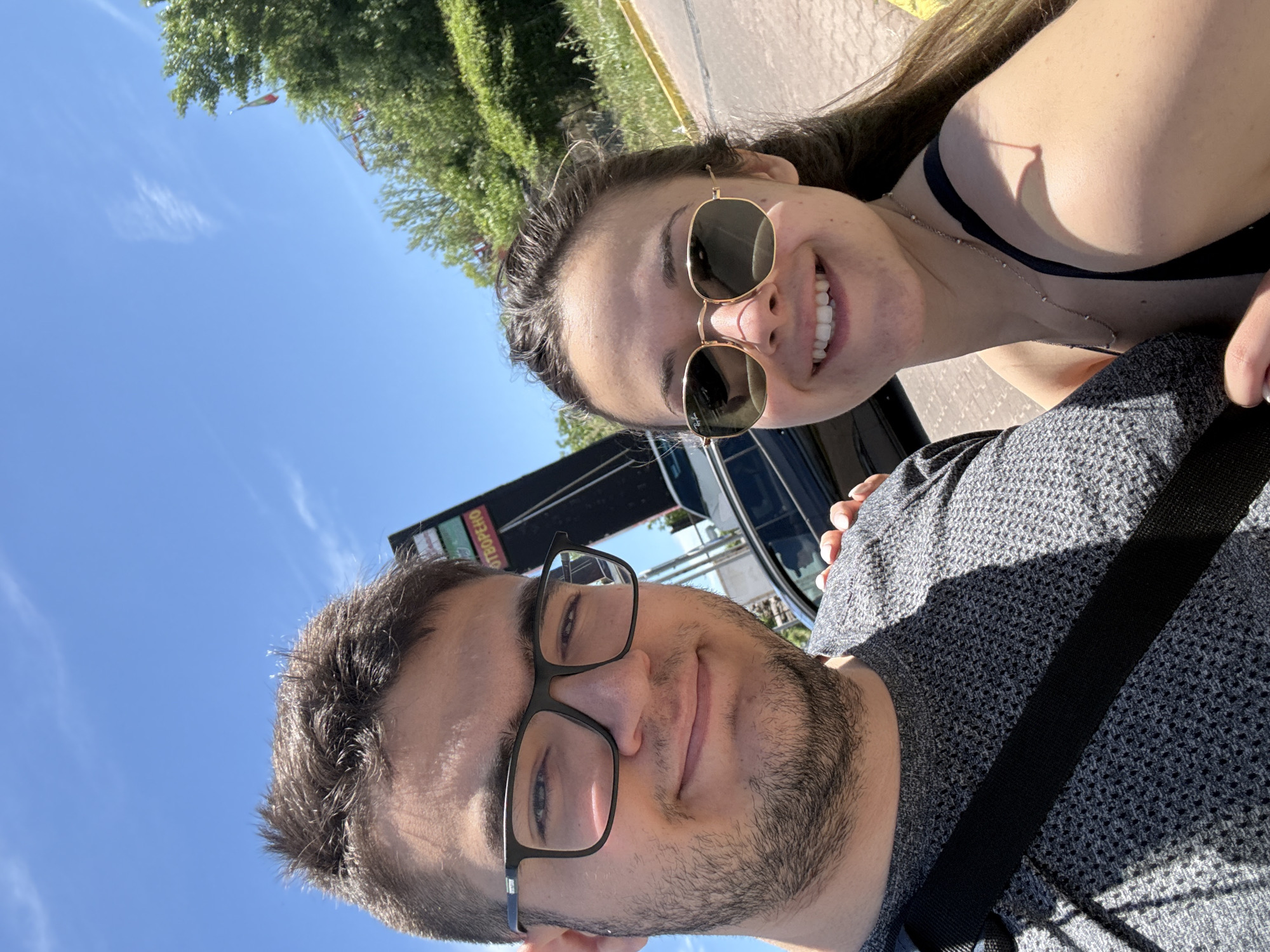 A selfie taken by a couple in a car in Sofia, Bulgaria. The woman is smiling and wearing sunglasses, with a dark top. The man is also smiling and wearing glasses and a gray sweater, with a seatbelt visible. Trees and the sky are visible in the background, as well as a bus and a building with signage in Cyrillic.