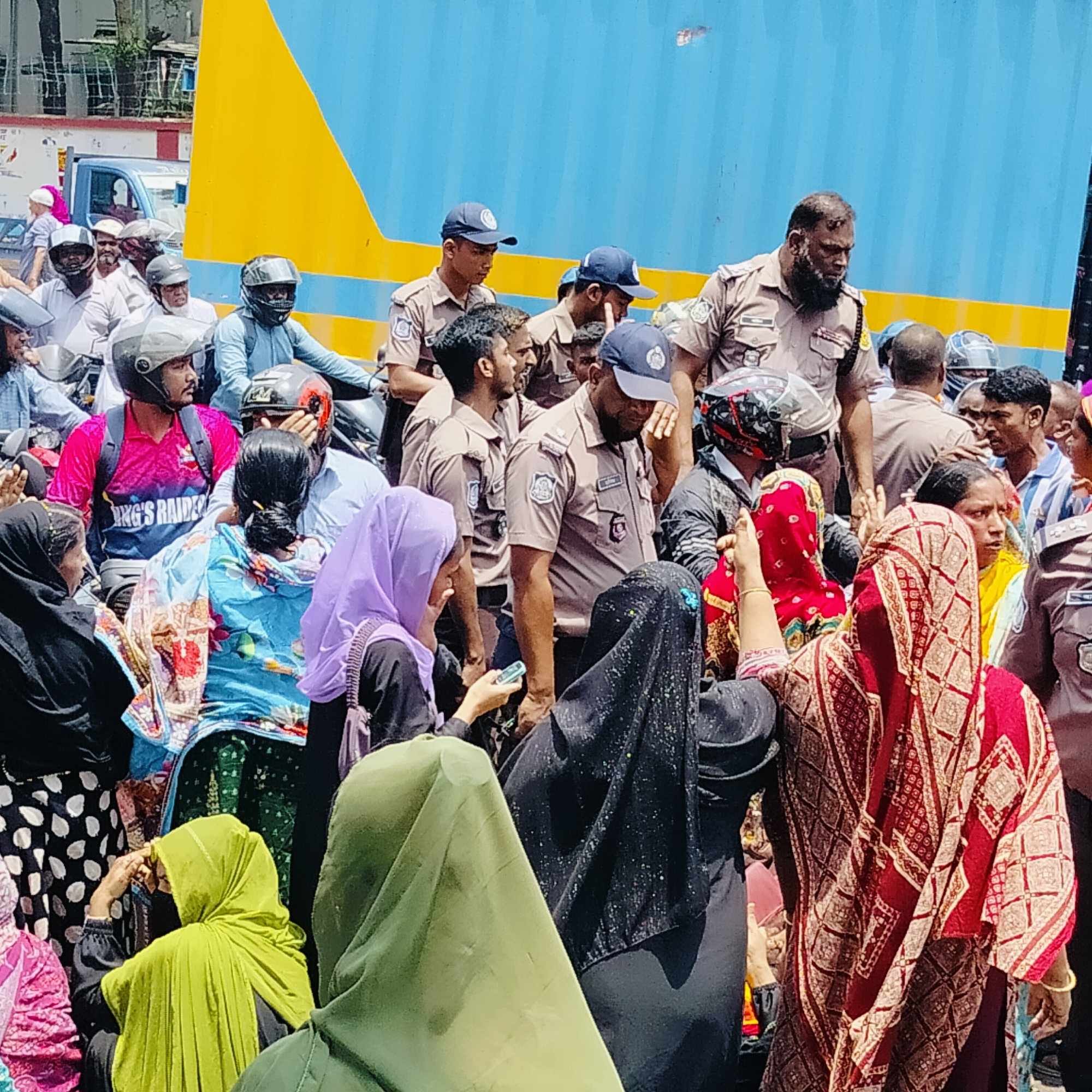 The image captures a crowded street scene in Dhaka, Bangladesh. A group of police officers in khaki uniforms and caps stand amidst a gathering of people. Many of the women in the crowd are wearing hijabs or burkas in a variety of colors. Motorcyclists wearing helmets are also visible in the background, along with a blue and yellow container truck. The scene appears to be outdoors on a bright, sunny day.