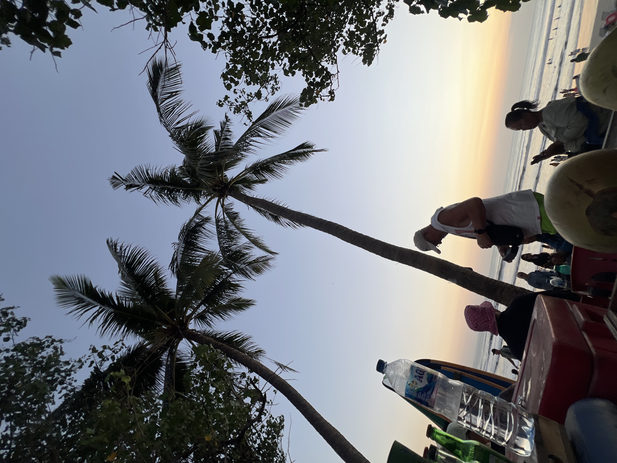 The image captures a sunset scene at Kuta Beach, Bali, Indonesia. In the foreground, a table holds a bottle of Aqua water, a red cooler, and some green bottles, with two large, light green, spherical objects partially visible. Two palm trees frame the sky, which transitions from a pale blue to a soft orange and yellow at the horizon.
Several people are present. A man in a white tank top and a baseball cap is in the mid-ground, holding a small black object. To his left, a person wearing a pink bucket hat is seated, facing away from the camera. Further back, other individuals are visible, some near the water's edge, where waves are breaking on the shore. A woman with her hair in a ponytail is seated on the right, looking towards the ocean. The overall atmosphere suggests a relaxed, tropical evening.