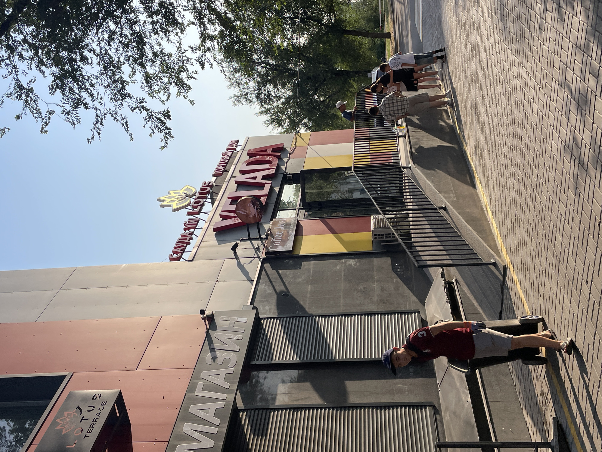 In Almaty, Kazakhstan, a street scene unfolds under a clear sky. A multi-story building with modern architecture, featuring red and gray paneling, serves as the backdrop. A prominent sign on the building displays "LOTUS TERRACE" with a lotus flower emblem. Below, a large sign reads "МАГАЗИН," indicating a shop. Further along the building, a red sign with a golden lotus emblem displays "VELADA," along with smaller text suggesting a shopping center and a "Coffee Jos" establishment.
Several people are present. A young man in a maroon polo shirt, gray shorts, a dark baseball cap, and sandals is seen pushing a large, wheeled trash bin on a paved walkway. To the right, a group of individuals, including adults and children, are gathered near the entrance of the "VELADA" establishment. One adult, wearing a plaid shirt and shorts, appears to be interacting with a child in a black dress and another child in a white and blue striped shirt and denim shorts. Another person, partially visible, is standing further back.
The environment includes a paved pedestrian area with a yellow dividing line, a metal railing, and a dark gray wall. Trees with green foliage are visible in the background, suggesting a well-maintained urban area. The sunlight casts distinct shadows, indicating it is likely late afternoon. The building's facade is accented with colorful panels in red, yellow, and white.