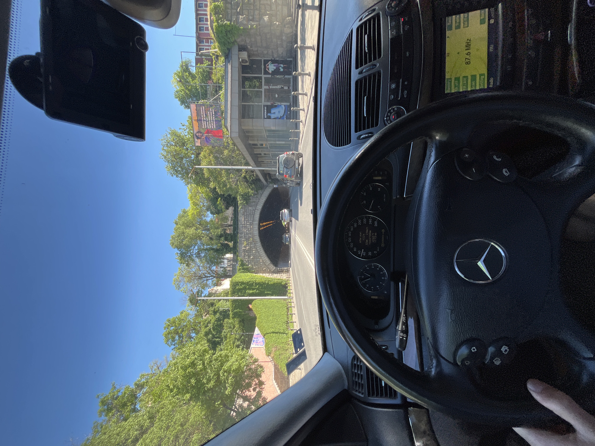 A view from inside a Mercedes-Benz E-Class, looking towards the Plovdiv underpass tunnel on Tsar Boris III Obedinitel Boulevard in Plovdiv, Bulgaria. The dashboard, steering wheel (with the Mercedes emblem), and part of the windshield are visible. Outside, the tunnel entrance is framed by trees and green hedges. A billboard and pedestrian traffic are present in the background. The radio frequency on the dashboard display is 87.6 MHz. The speedometer indicates the car is stopped with 117.5km on the odometer.