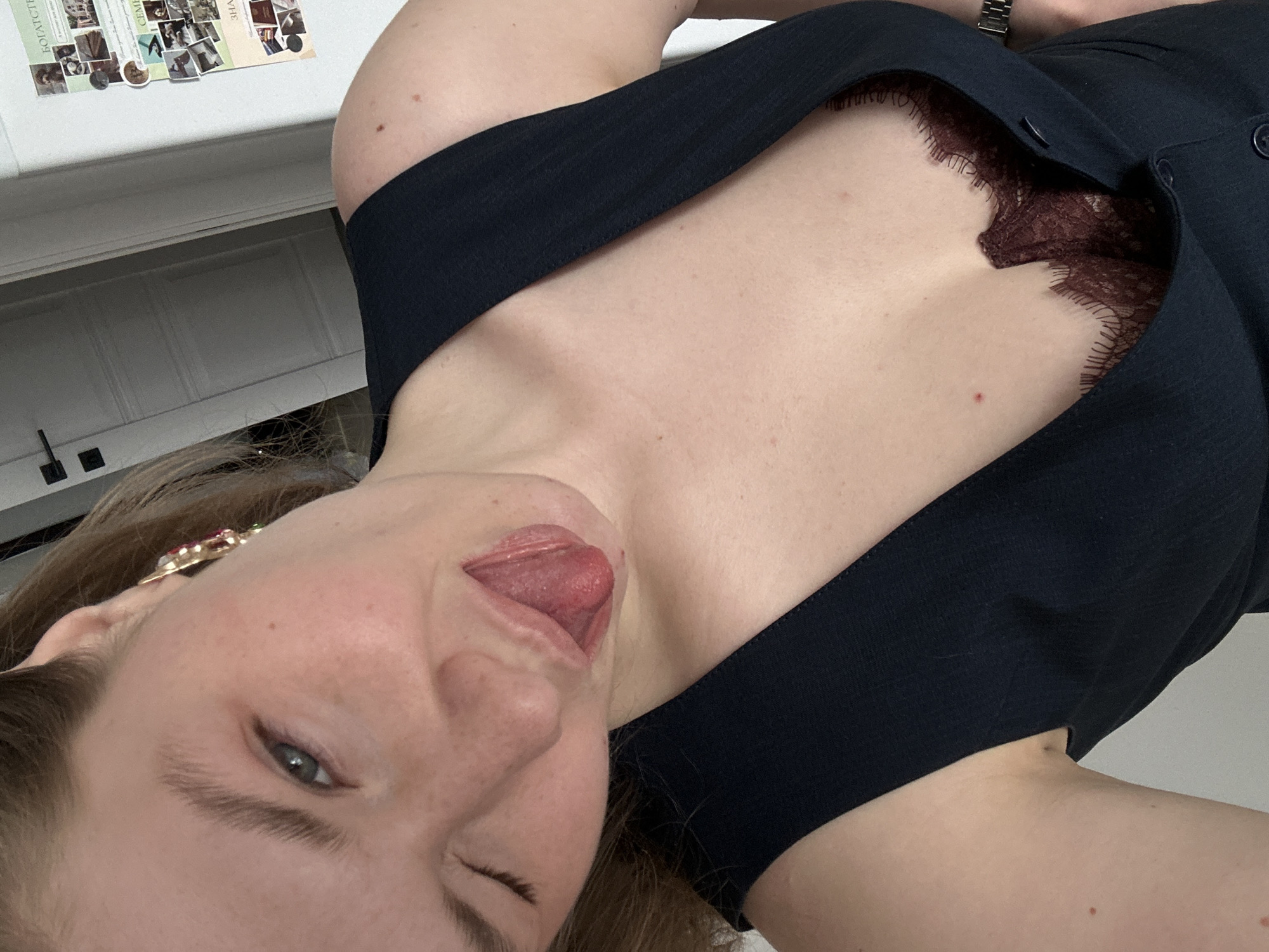 A close-up, angled selfie depicts a woman with fair skin sticking out her tongue and winking one eye. She has light brown hair and is wearing a black, low-cut garment that reveals a glimpse of burgundy lace underneath. A decorative hairpin is visible in her hair near her left ear. The background features a light-colored wall and a partial view of a bulletin board adorned with photos. A kitchen cabinet can be seen in the background.
There is nothing in the image that indicates it was taken in Kazan, Russia.
