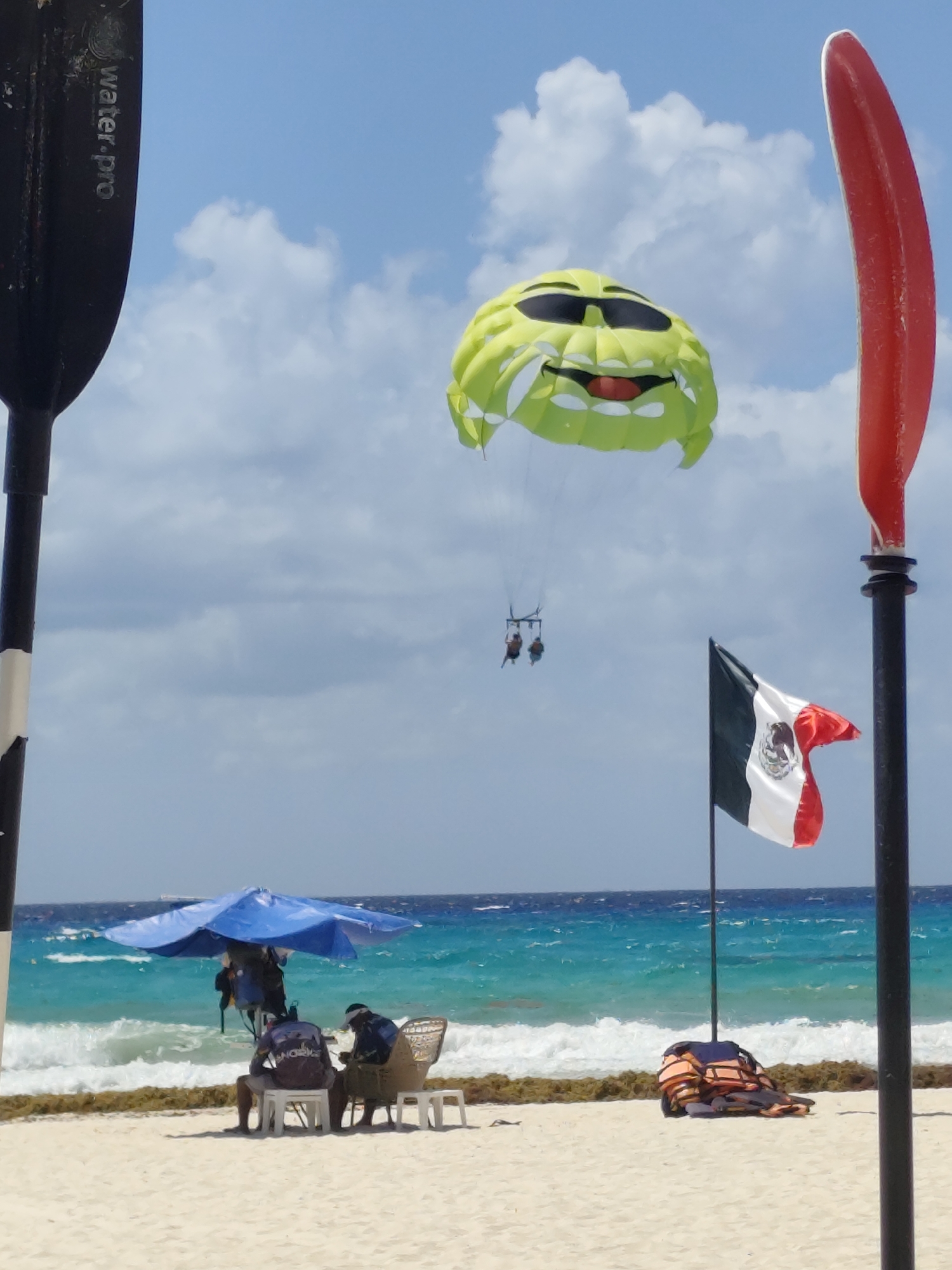 The image depicts a beach scene in Playa del Carmen, Mexico, characterized by a bright, sunny day. A green parasail with a cartoon face wearing sunglasses soars in the sky above the ocean, carrying two people in a harness below. In the foreground, the left side of a black and white paddle is seen, along with a red paddle on the right, partially obstructing the view. The Mexican flag is placed to the right of the image's center, fluttering in the wind. On the beach, a cluster of life jackets sits in front of the flag. Further to the left, under a blue umbrella, two individuals sit on plastic chairs, potentially beach service personnel. The ocean is a mix of blue and green, with breaking waves and seaweed near the shore. The sand is light-colored and occupies the bottom third of the frame.
