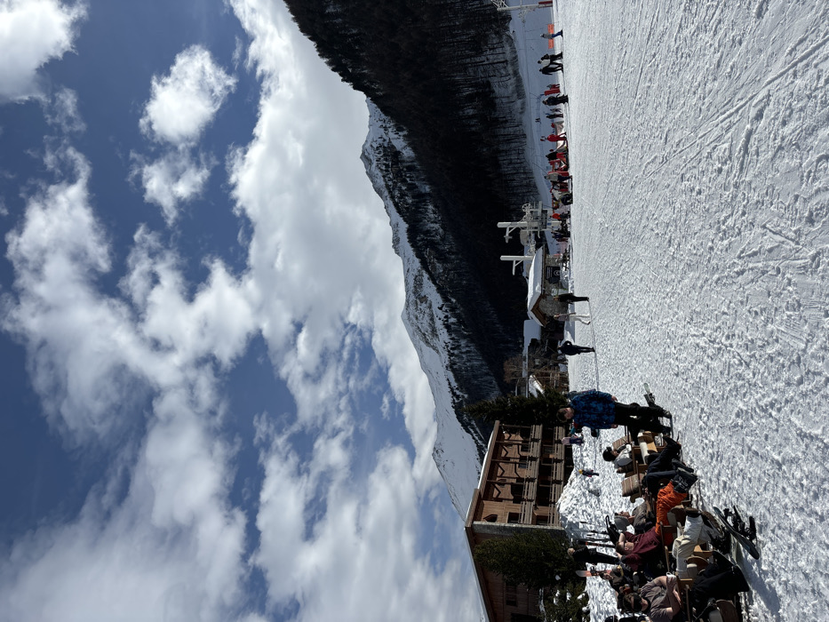 This image depicts a vibrant outdoor scene at a ski resort, likely Val-d'Isère, France, under a clear blue sky dotted with scattered white cumulus clouds. The photograph, originally captured in a portrait orientation but showing a landscape scene, reveals a broad ski slope filled with tracks, leading up towards snow-capped mountains and a dark, dense forest in the background.
In the mid-ground, at the base of the slope, are resort buildings and infrastructure. Notably, a prominent white sign with black lettering reads "LA FOLIE DOUCE," identifying a famous après-ski establishment. Next to it are other white structures, possibly related to ski lift operations.
The foreground is bustling with activity, showing numerous people gathered at an outdoor seating area. Many individuals, dressed in colorful ski jackets, pants, and helmets, are either seated at tables or standing, socializing and relaxing. Skis and poles are visible, propped against tables or lying in the snow. A multi-story wooden lodge-style building is situated on the left side of this gathering area. The time of day appears to be midday or early afternoon, with bright sunshine casting distinct shadows, indicating good weather conditions. The overall atmosphere is relaxed and social, suggesting a break from skiing or an après-ski event.