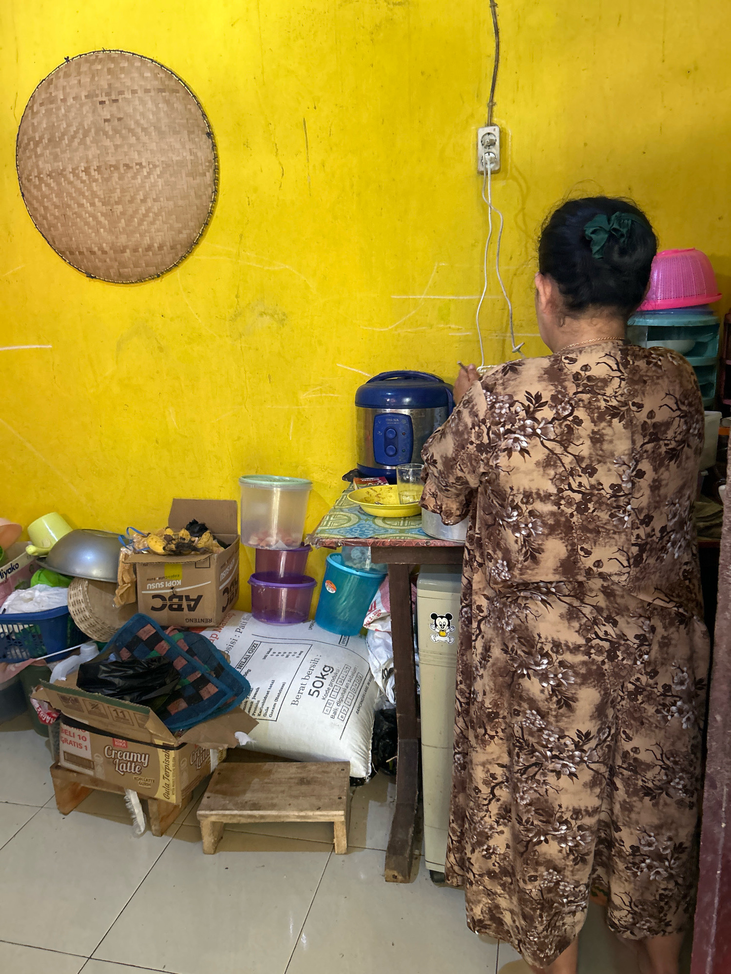 The image shows a woman wearing a brown floral dress standing in a room with yellow walls. She is facing away from the camera, towards a rice cooker and various kitchen items. The room appears to be a storage area or kitchen, with stacked plastic containers, a large bag of rice, boxes, and other household goods. There is a woven mat hanging on the wall, and a power outlet with wires. The floor is tiled. The overall impression is of a cluttered, lived-in space.