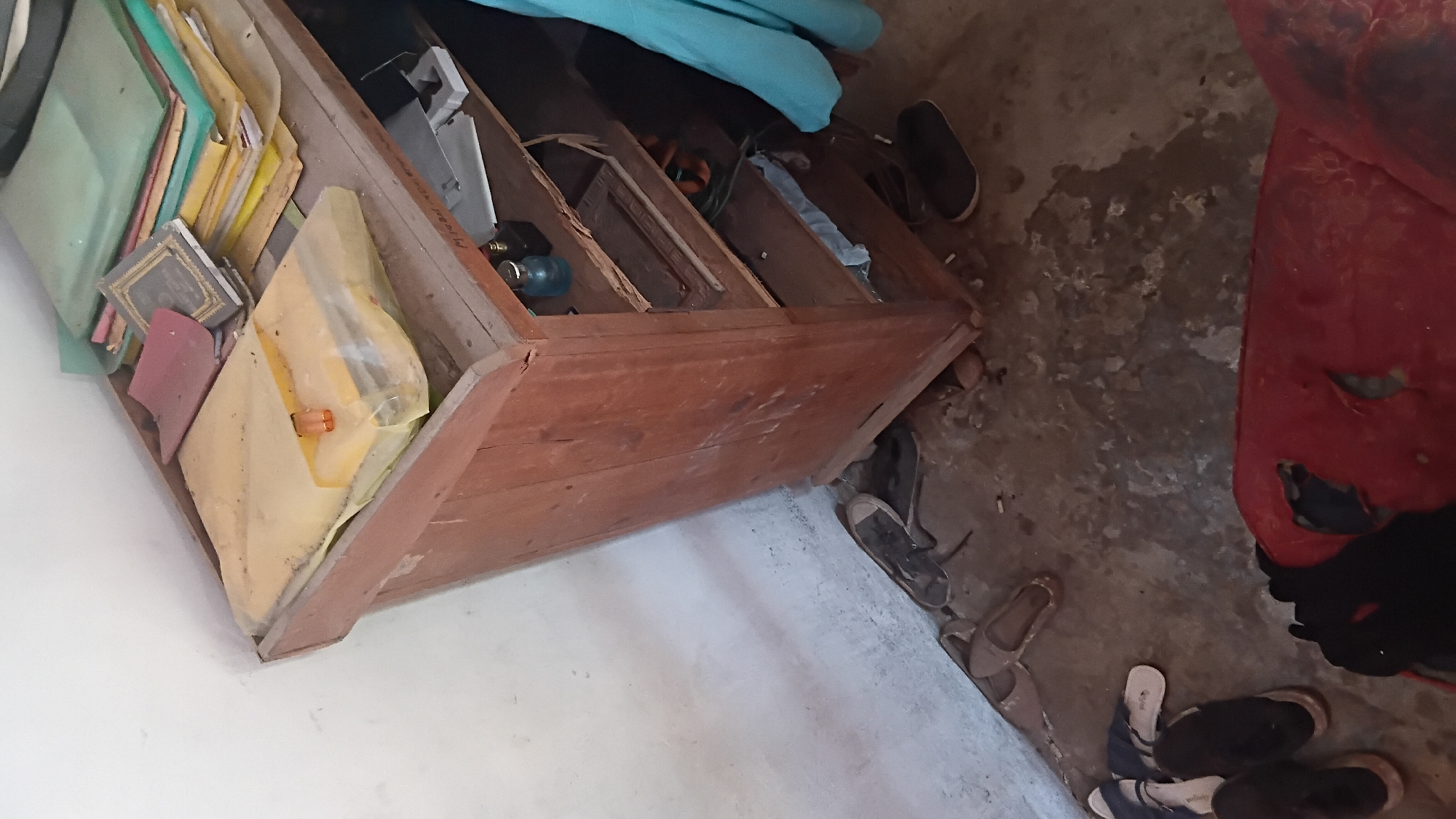 The image depicts an indoor scene in Prabumulih, Indonesia, featuring a cluttered wooden cabinet and the surrounding floor area. The cabinet, made of aged wood, has shelves filled with various items, including stacks of documents and books. Some items are covered with plastic sheeting. On the top shelf, there are two small bottles, one blue and one dark. On the floor surrounding the cabinet, several pairs of footwear are visible, including sandals and shoes. A portion of a red, damaged cushion or piece of furniture is also present in the foreground. The environment appears somewhat neglected, with dust evident on the surfaces. The lighting suggests an indoor setting, possibly a storage room or a poorly maintained living space.