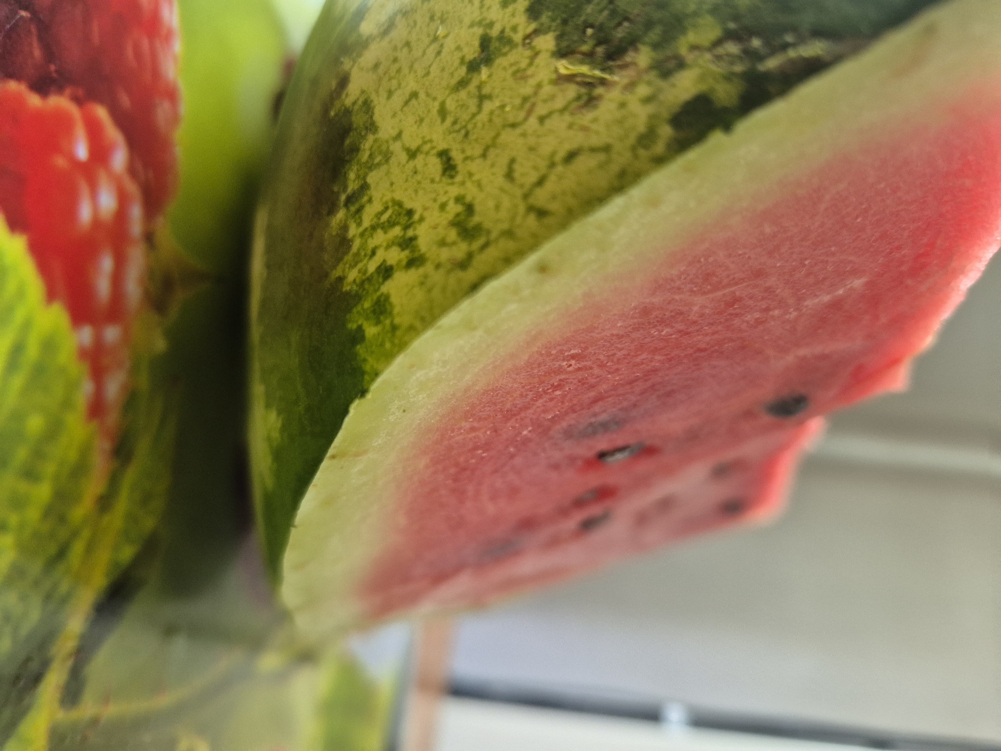 A close-up shot focuses on a slice of watermelon, showcasing its vibrant red flesh and green rind. The watermelon appears to be resting on a surface decorated with images of raspberries. The background is blurred, but hints of a white wall and possibly a window are discernible. The lighting suggests an indoor setting, possibly a kitchen or dining area. There is no indication of the location within the city of Kokshetau, Kazakhstan.