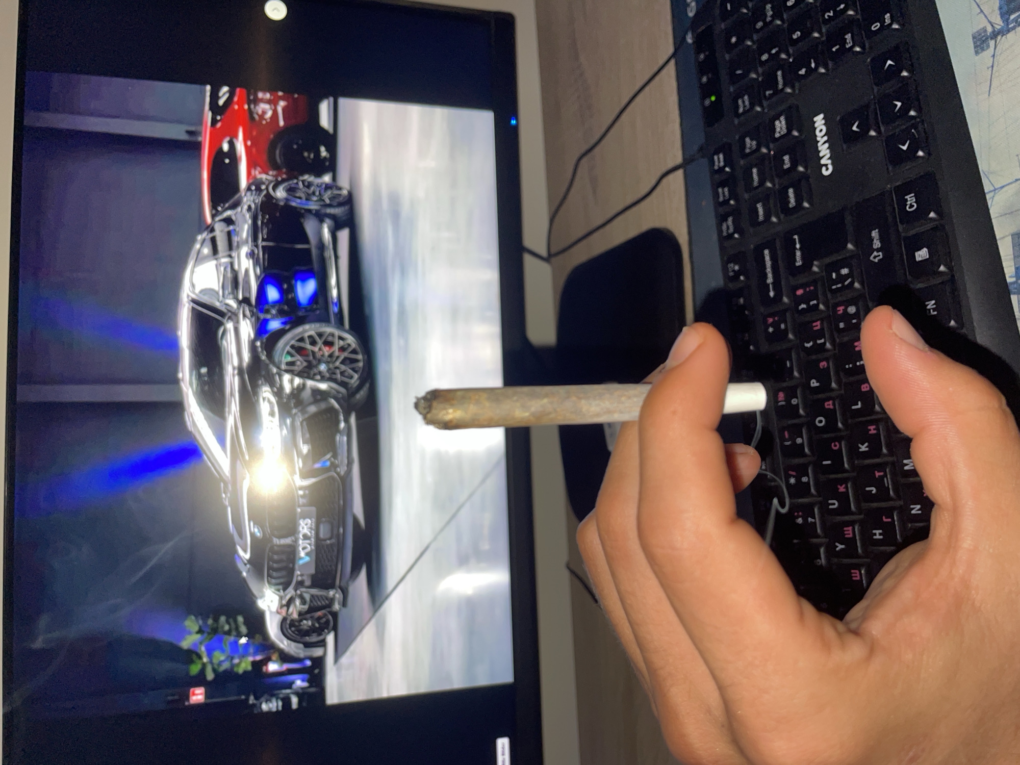 The image depicts an indoor scene with a close-up on a hand holding a lit, hand-rolled item over a computer keyboard, with a monitor displaying an image in the background. The location is Unknown, Unknown.
In the foreground, a light-skinned hand, likely belonging to an adult, is holding a partially consumed, hand-rolled cylindrical item between the index finger and thumb of the right hand. The tip of the item is charred, indicating it has been lit and smoked, while the lower part held by the fingers has a white filter or crutch. Beneath the hand, a black computer keyboard with both Latin and Cyrillic character keycaps is visible. The brand name "CANYON" is prominently displayed on the keyboard. A black cable runs from the keyboard towards the back of the desk.
In the mid-ground, a flat-screen computer monitor displays an image of several luxury cars in what appears to be a showroom or exhibition space. The central and most prominent car is a dark metallic BMW, possibly an M series coupe SUV, with bright headlights and a large front grille. A blue-lit logo reading "V MOTORS" is visible on its front bumper. To the left of the central car, a lighter-colored vehicle is partially visible, while to the right, a red SUV or crossover is partially obscured. The cars are positioned on a reflective, light-colored floor, against a dark background illuminated by distinct blue spotlights or light beams. Hints of green foliage are visible on the far left of the displayed image. The monitor rests on a black stand, situated on a light-colored wooden or laminate desk surface.