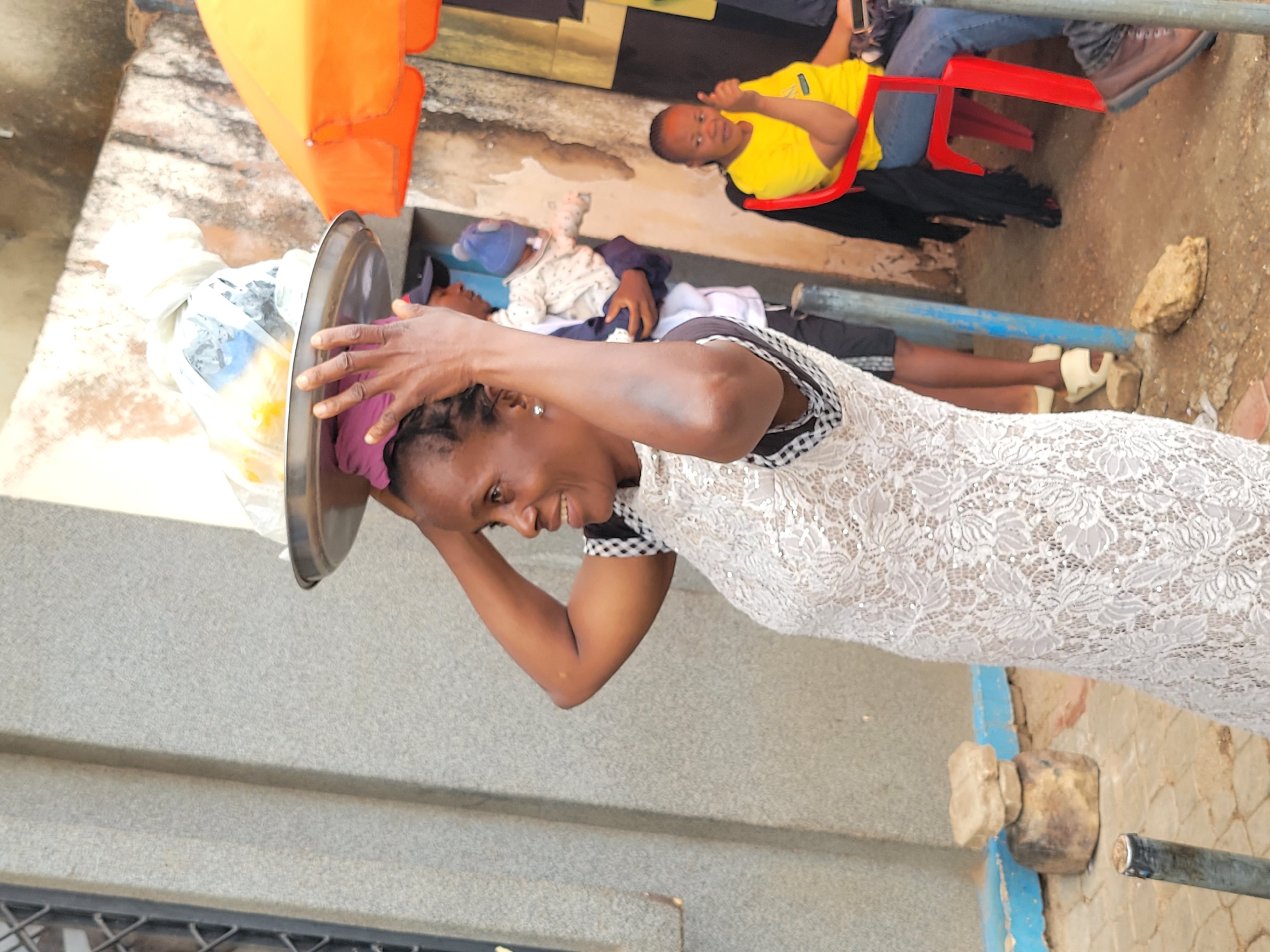 The image shows a woman in Jos, Nigeria, balancing a tray on her head. The woman, wearing a white lace dress, is smiling and steadying the tray with her hand. The tray contains items wrapped in plastic bags. In the background, there are two individuals; one is wearing a yellow shirt and sitting on a red chair, the other is lying down. A light-orange tarp is visible. The environment is a street scene with a mix of concrete and weathered surfaces.