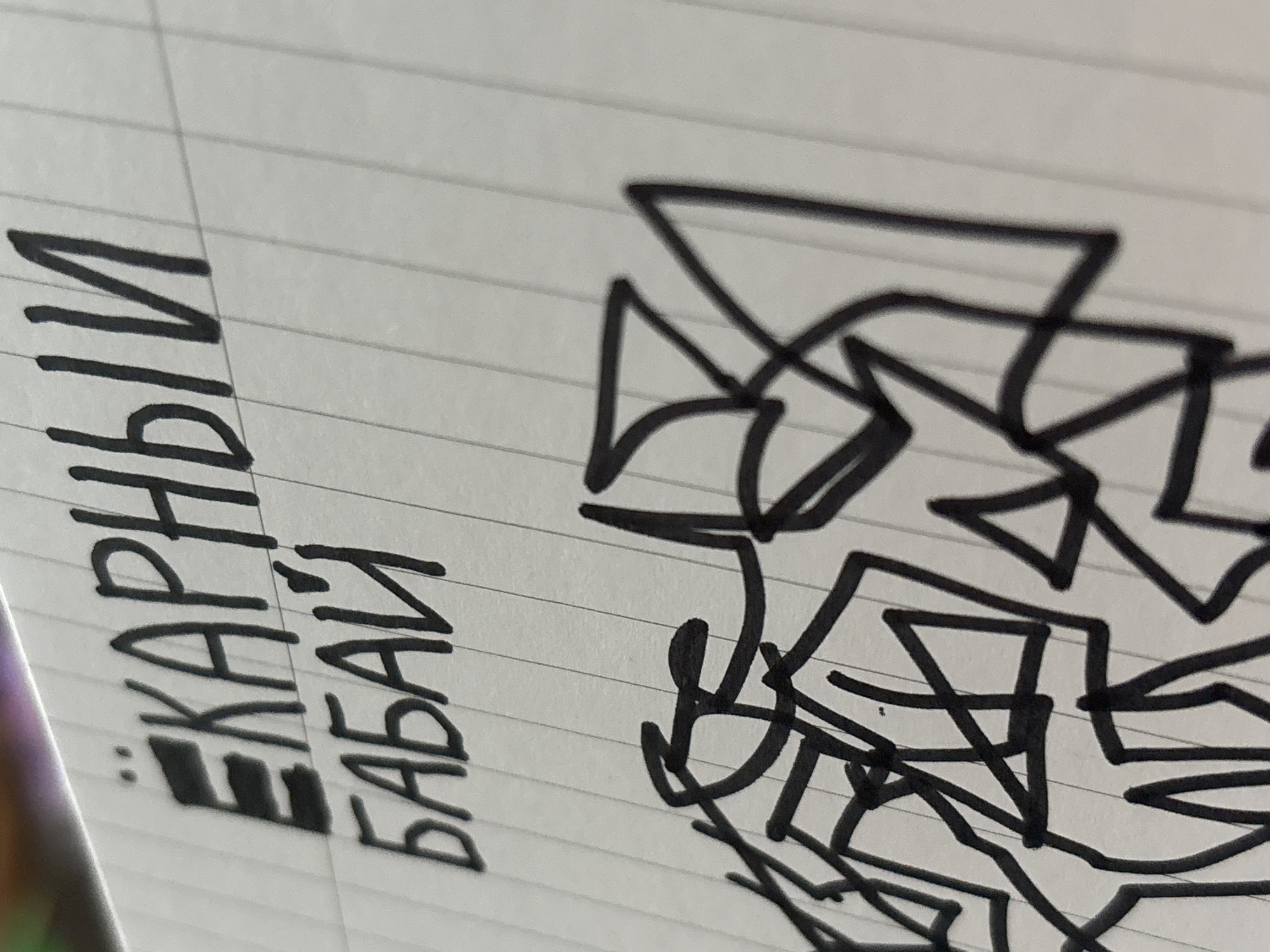 The image shows a close-up of a piece of lined paper with text and a drawing. The text is written in black marker and reads "ЕКАРНЫЙ БАБАЙ". Below the text is a drawing made with thick black lines, appearing to be abstract or graffiti-style art. The background is a lined notebook page, with horizontal lines visible throughout. There is no indication of the location in Saint Petersburg, Russia.