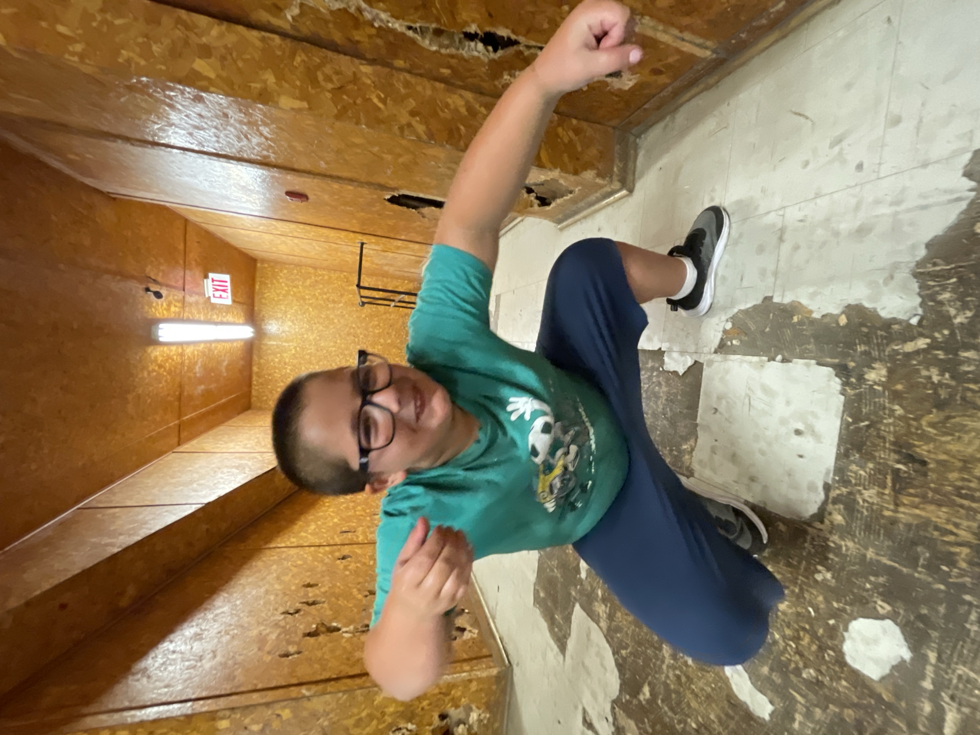 A young male wearing glasses, a green t-shirt, and dark blue pants is crouching in a hallway. The hallway has wood-paneled walls and a drop ceiling with fluorescent lighting. An "EXIT" sign is visible on the wall. The floor appears to be tiled and in disrepair, with some tiles missing and others cracked. The boy's left arm is extended forward, and his right fist is clenched. He is looking at the camera with a smile. The scene does not provide specific details to determine its location in the city of Bluewater, Canada.
