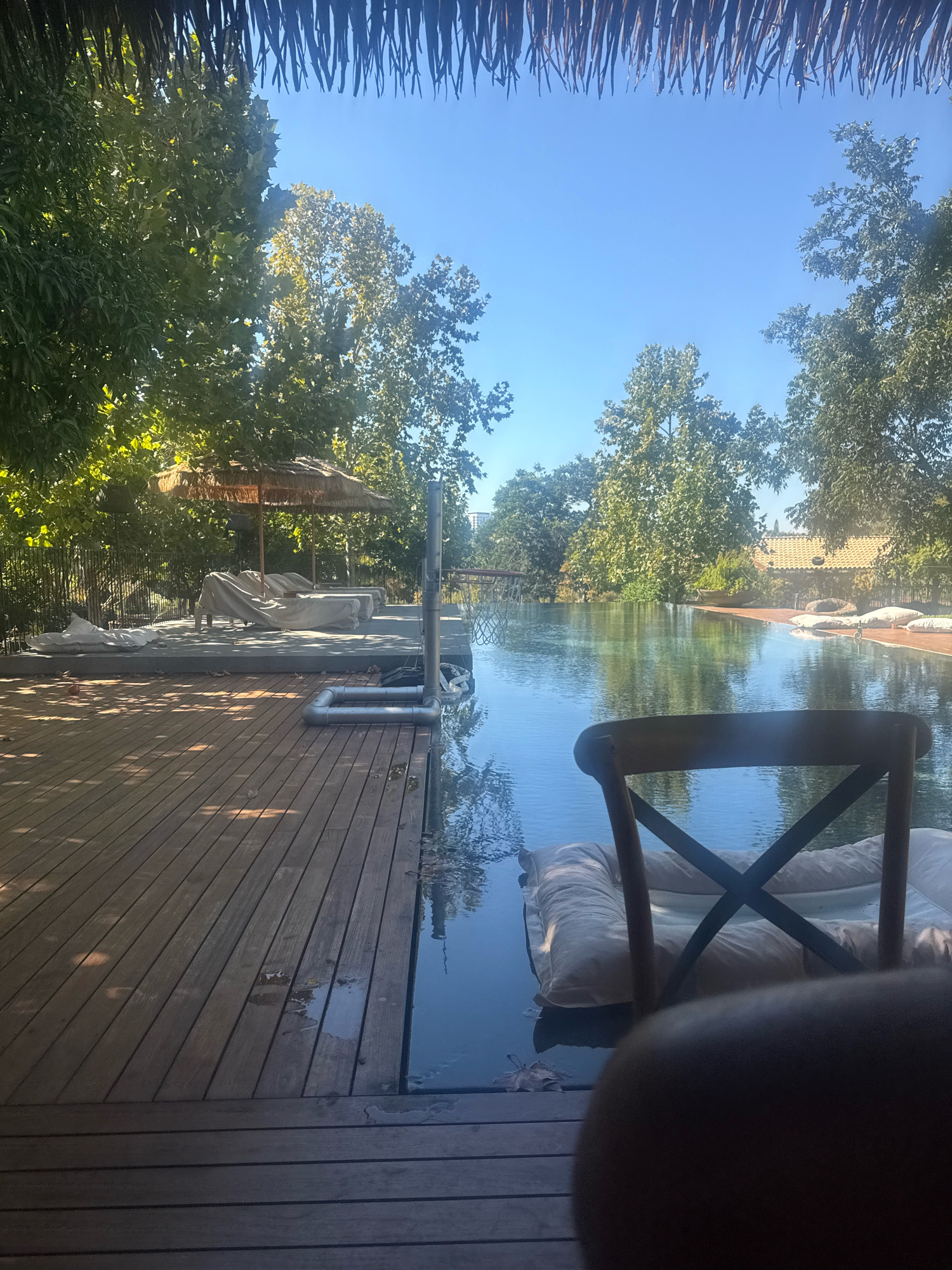 The image depicts an outdoor scene, likely at a resort or a private property in Drom HaSharon Regional Council, Israel. A wooden deck leads to a large, tranquil swimming pool with an infinity edge. The pool is surrounded by lush green trees and a clear blue sky.
In the foreground, a wooden chair is partially visible, facing the pool. A large, cream-colored inflatable lounger floats in the water, positioned in front of the chair. The deck is made of dark wooden planks, with some leaves scattered on its surface, suggesting an autumn setting or natural debris from the surrounding trees.
To the left of the pool, there's a concrete platform with two lounge chairs covered in light grey fabric, and a thatched umbrella providing shade. A black metal fence runs behind this area. Various pipes and plumbing fixtures are visible near the edge of the pool, indicating the pool's infrastructure.
In the distance, across the pool, a tiled roof of a building is visible, along with more greenery. The overall atmosphere is serene and relaxing, with the sun casting dappled shadows on the wooden deck. The image appears to be taken during daylight hours.