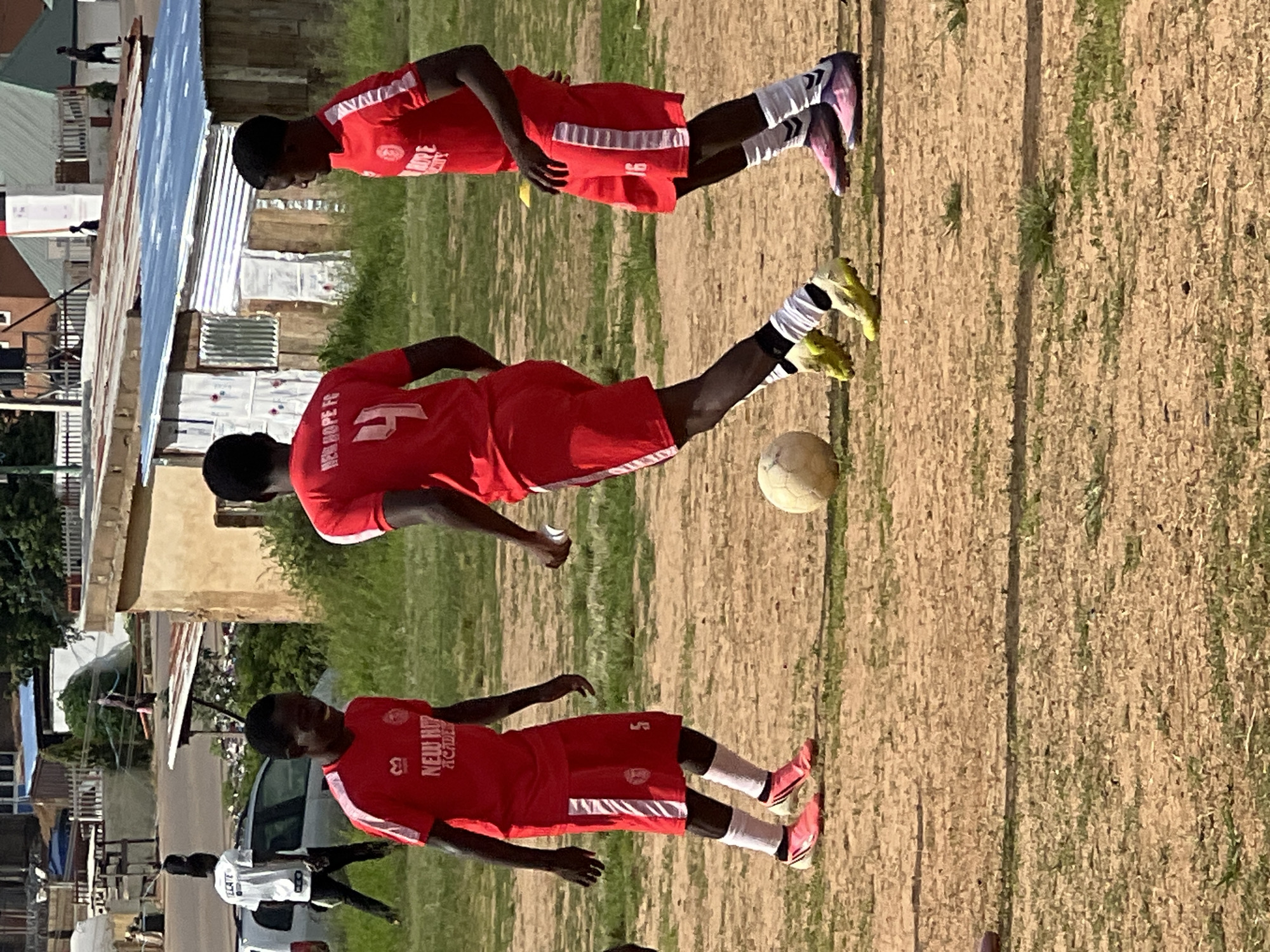 In Jos, Nigeria, three young men in red soccer uniforms with white trim are playing soccer on a dirt field with sparse grass. Two of the players are wearing white socks, and the third is wearing white and black striped socks. One player has the number 4 on his jersey and another has the number 16. The player on the left is wearing shorts with the number 5. A soccer ball is visible on the ground. In the background, there are buildings with corrugated metal roofs, a car, and a small group of people standing near a fence. The sky is visible and appears to be clear.