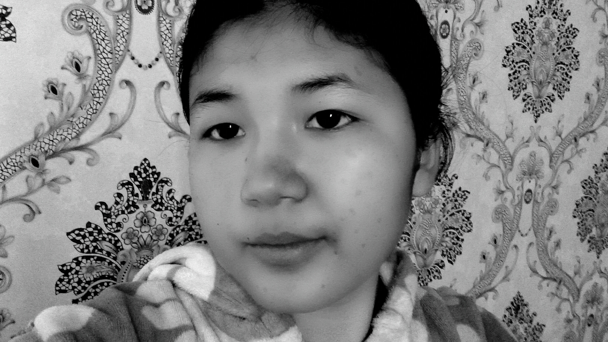 A black and white, medium-close shot shows a young East Asian person from the chest up, facing the camera. They have dark hair pulled back, dark eyes, and are wearing a patterned hooded garment that appears to be a robe. The background is a repeating floral wallpaper pattern. The lighting casts a slight shadow on the left side of their face. The location is stated as Uchkun, Kyrgyzstan, though no specific landmarks or environmental details are visible in the image to confirm this.