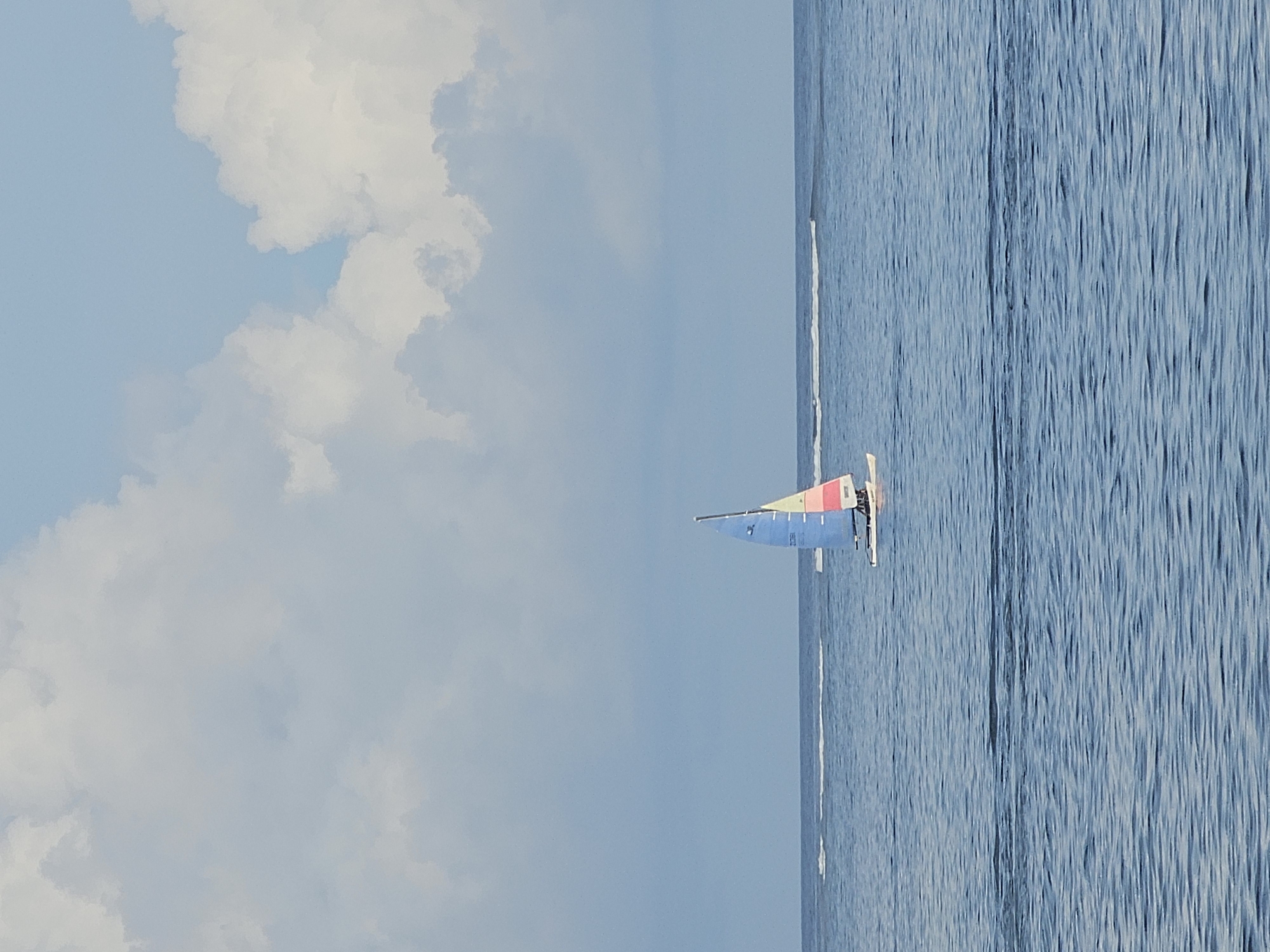 A sailboat is sailing on the sea under a blue sky with white clouds. The water has a visible ripple pattern. The photograph appears to be taken during the daytime. There is no indication of this photo being taken in Denpasar, Indonesia.