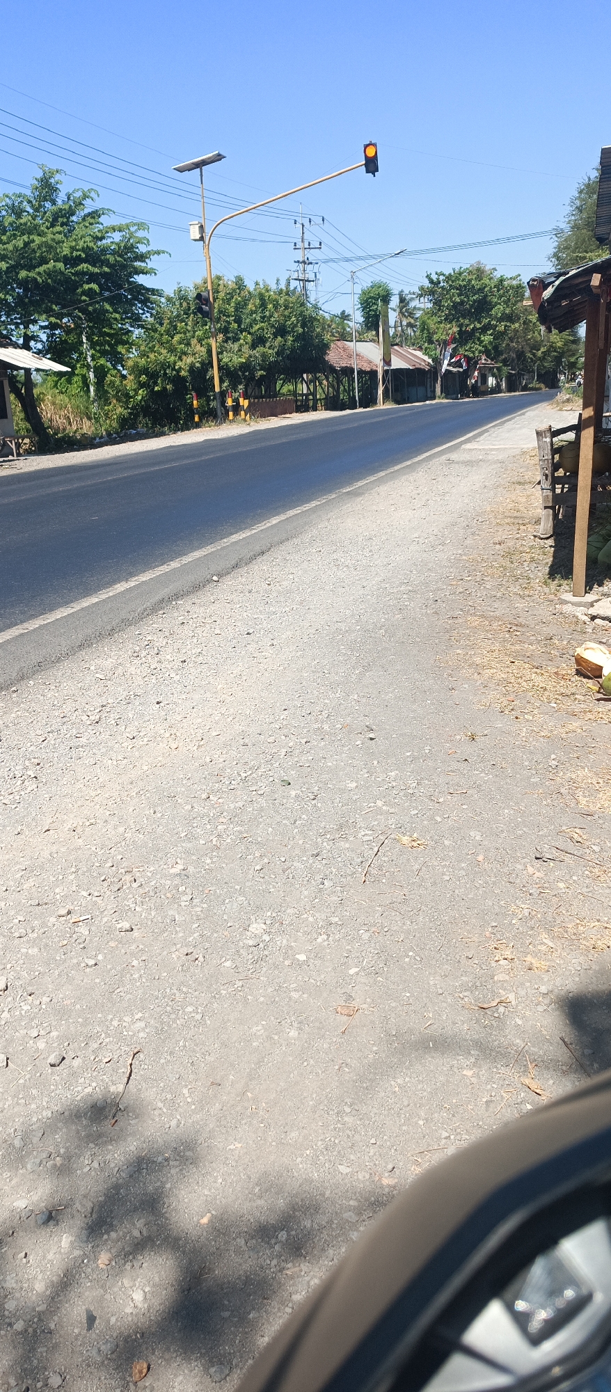 A low-angle, eye-level shot captures a sunny day on a paved road in Jatisari, Indonesia. The road surface is a mix of asphalt and gravel. On the left side of the road, lush green trees line the verge. A traffic light with an amber signal hangs over the road, supported by a pole equipped with a solar panel. On the right side of the road, a wooden structure with a thatched roof stands, and several coconuts are visible on the ground. The sky is a clear, bright blue. The image appears to have been taken from a vehicle.