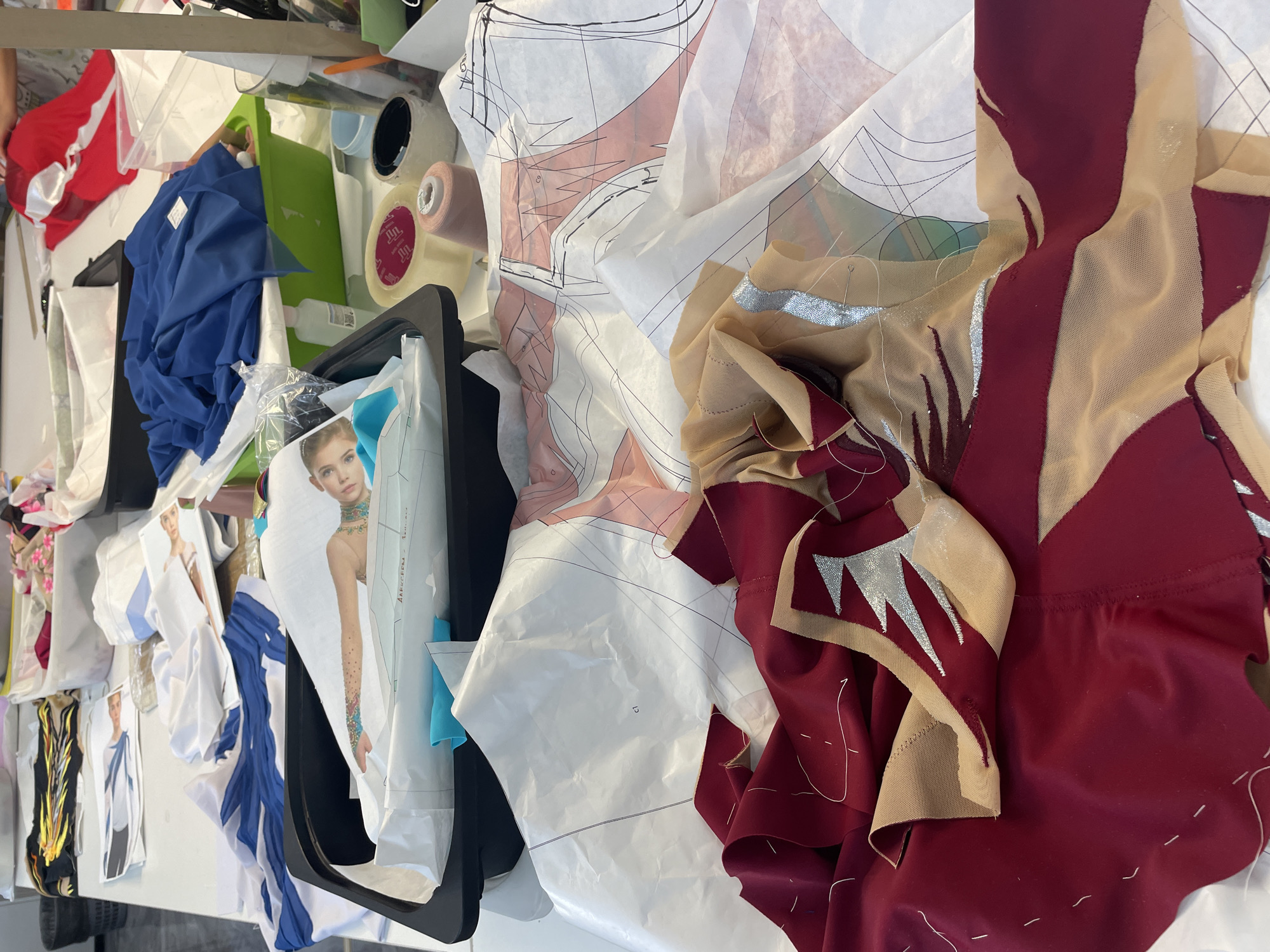 This is a close-up, overhead shot of a table laden with materials for costume making, likely in a workshop or studio. The scene is dominated by fabrics, pattern paper, and various sewing supplies.
In the foreground, a deep red fabric is draped over beige mesh material. The red fabric is detailed with sharp, angular designs in a metallic silver or gold color, creating a striking contrast with the base colors. Some of the fabric appears to be loosely pinned, indicating it's in the process of being worked on. The pattern paper underneath is covered in intricate lines and measurements, typical of sewing patterns.
Further back, a black plastic tray holds a sheet of paper with a printed image of a person adorned with jewelry and a colorful, tattooed arm. This is likely a design or inspiration for the costume. Beside this tray, there's a roll of clear tape and spools of thread in various colors, including a pale pink.
To the right, a bright blue fabric is folded and placed inside a green plastic container, suggesting it's a material waiting to be used. In the background, more fabric and supplies are visible, including a white container with what looks like red and white fabric.
The overall impression is one of creative activity and preparation for a crafting project. The lighting is bright and even, illuminating the details of the materials and the workspace. The specific location in Saint Petersburg, Russia, is not discernible from the visual information provided in the image.