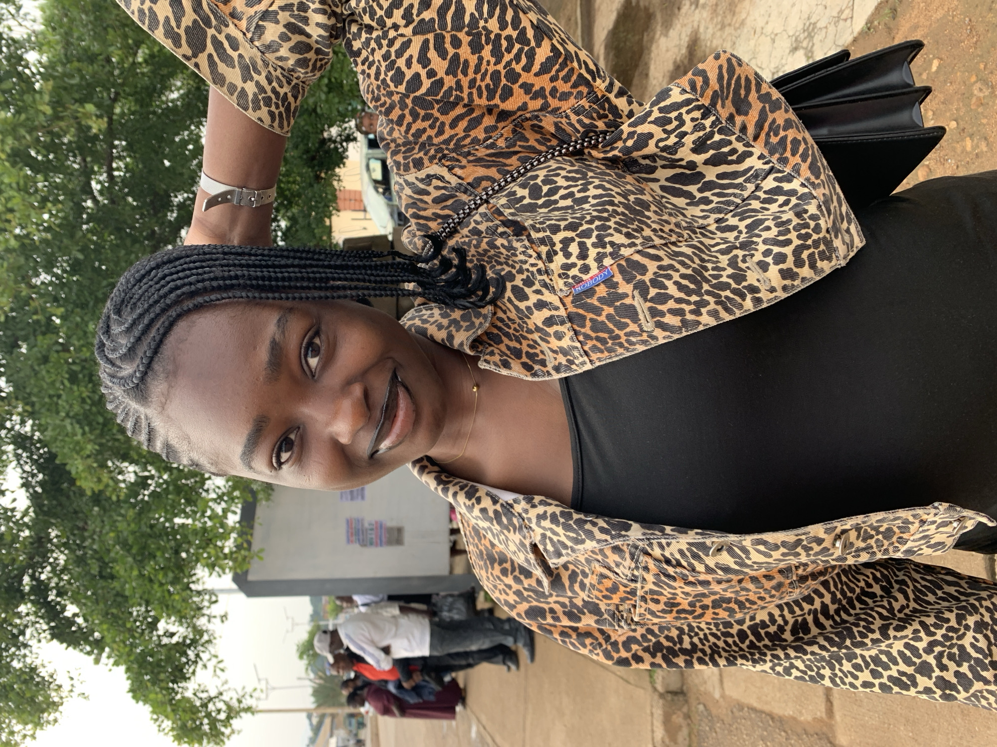 The image shows a young Black woman in Rafin Par, Nigeria, standing outdoors. She is wearing a black tank top and a leopard print denim jacket. Her hair is styled in braids, and she is wearing dark lipstick and a delicate gold necklace. A silver watch is visible on her right wrist. She is holding her right arm up, resting her hand behind her head, and looking at the camera with a smile. A black handbag is visible on her hip. In the background, several people are standing on a paved area next to a building, with trees and a road visible further in the distance.