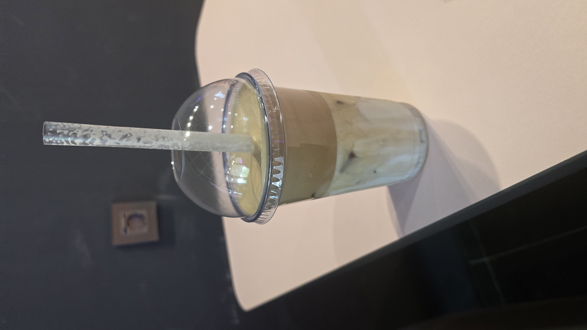 The image displays a clear plastic cup filled with a layered beverage, likely a coffee drink, topped with a domed plastic lid. A clear, textured plastic straw extends from the lid into the drink. The beverage itself exhibits distinct layers, with a lighter creamy color at the bottom, a darker brown layer in the middle, and a lighter, foamy layer at the top. Ice cubes are visible within the drink. The cup is positioned on a surface that appears to be a table or countertop, with a dark, reflective surface in the foreground and a lighter, possibly paper or fabric, surface behind the cup. The background on the left is a dark, textured wall with a small, framed picture. The lighting suggests an indoor setting, possibly a cafe or home. No specific time of day or weather conditions can be determined. The image is a close-up of the drink, with no people or specific activities explicitly shown. There is no visible text in the image. The provided location context of Ukhta, Russia, does not offer any direct visual cues within the image itself.