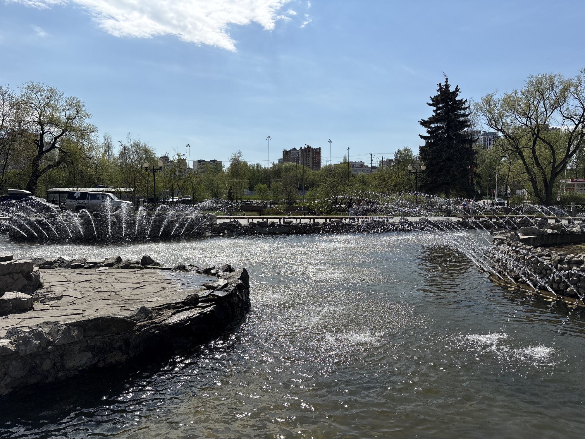 The image captures a water fountain in the city of Perm, Russia. The fountain is made up of multiple arcs of water jets spraying into a pond. Stone formations line the perimeter of the pond. In the background, there are trees, a tall evergreen, and buildings under a blue sky with sparse clouds. A car and people can be seen in the background amidst the park.