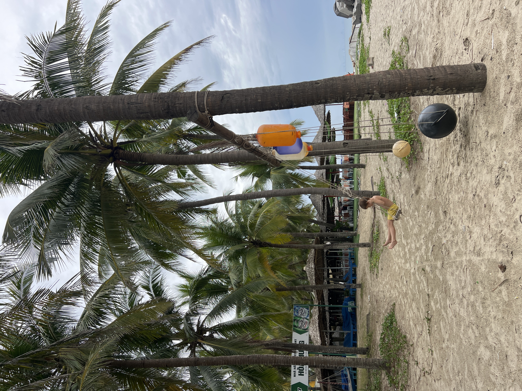 A young boy in yellow shorts is swinging on a swing in front of a row of thatched-roof huts and palm trees, on a sandy beach in Hoi An, Vietnam. The swing is suspended from a branch between two palm trees, with colorful buoys hanging nearby. In the background, the ocean is visible under a cloudy sky. The huts appear to be part of a restaurant or resort.