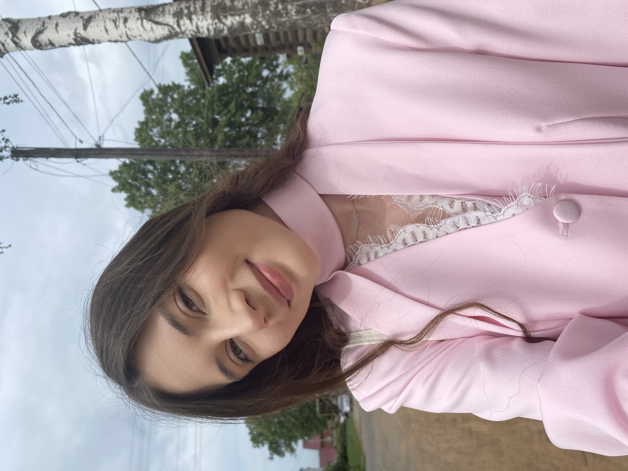 A young woman with long brown hair is wearing a pink jacket with a white lace undershirt. The photo was taken outdoors in Votkinsk, Russia, as evidenced by the birch tree and typical Russian buildings in the background. The sky is overcast. There are power lines visible above. The woman is looking directly at the camera with a slight smile.