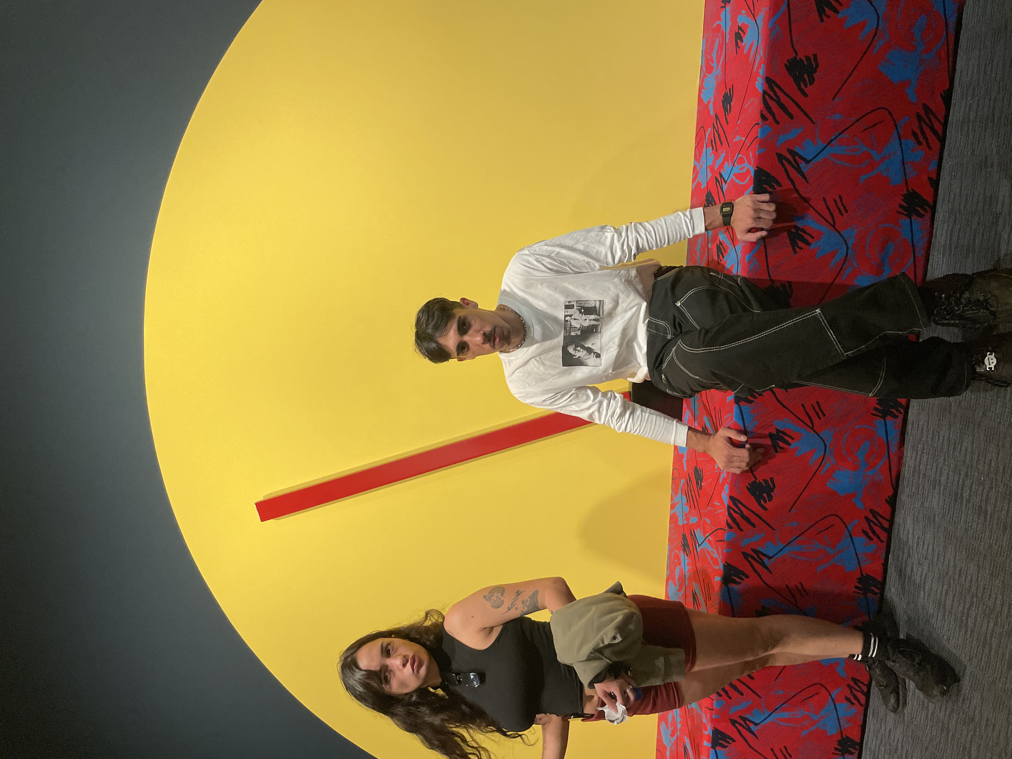 Two individuals, a man and a woman, are indoors in what appears to be an art installation or exhibit. The background features a large yellow circle with a red line extending from the center, resembling an abstract clock face. Adjacent to the yellow circle is a surface with a bold red, blue, and black patterned fabric. The floor is covered with gray carpeting.
The man is wearing a white long-sleeved shirt with a graphic on the front, black pants with white stitching, and dark shoes. He is positioned higher in the frame and appears to be leaning or sitting against the patterned surface.
The woman is wearing a black top, maroon shorts, and dark shoes with striped socks. She is holding a light green jacket or garment in one hand and some tissue in the other. The location is potentially an exhibit within the city of Buenos Aires, Argentina, but there is no clear evidence to confirm this detail from the image alone.