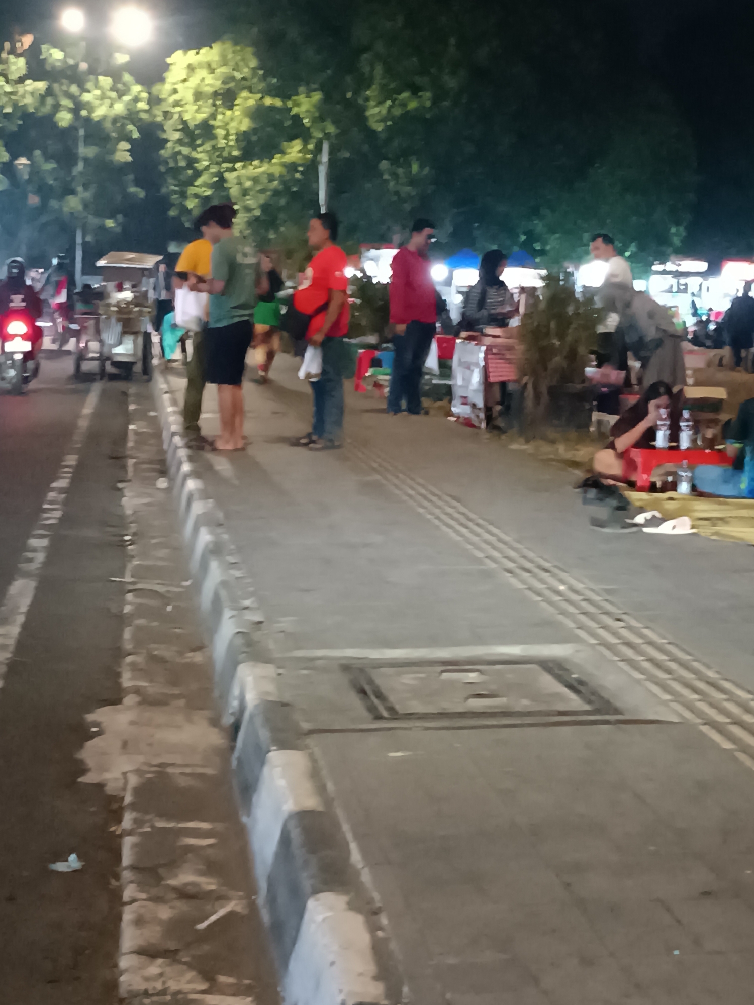 The image depicts a street scene at night in Yogyakarta, Indonesia, likely a pedestrian walkway or sidewalk adjacent to a road. Several individuals are visible, some standing and others seated. In the foreground, a paved sidewalk runs alongside a road, marked with white lines. A drainage grate is present on the sidewalk. Further back, people are gathered around food stalls or vendor carts, suggesting a night market or street food area. Trees line the background, illuminated by overhead lighting. One person on a motorcycle is visible on the road to the left. The atmosphere appears casual, with people interacting and purchasing food. The lighting is a mix of streetlights and lights from the vendor stalls, creating a somewhat dim and atmospheric scene.