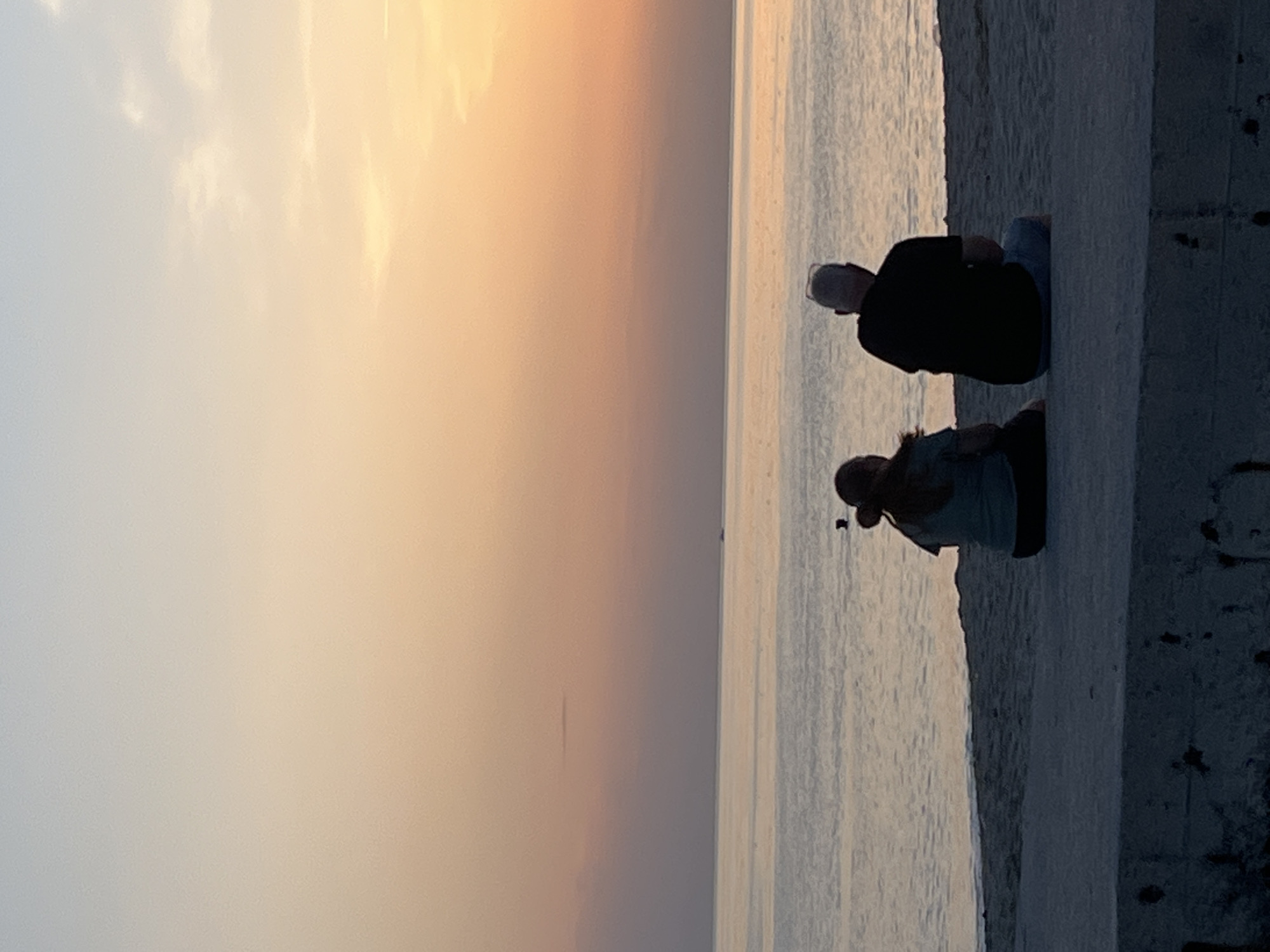 The image, oriented vertically, captures a tranquil waterfront scene at either sunrise or sunset in Alanya, Turkey. Two individuals are the main subjects, seen in profile, sitting or kneeling on a concrete structure that forms a barrier beside a calm body of water, likely the sea.
The person in the foreground wears a lighter-colored top, possibly blue or green, and darker bottoms, with their hair possibly tied back. The person further back wears a dark (likely black) long-sleeved shirt and light-colored pants or shorts, with what appears to be a clear visor or glasses on their head. Both are facing away from the camera, looking out towards the expansive horizon.
The setting is characterized by a soft, warm gradient sky, transitioning from a warm orange-pink hue near the horizon to a lighter, cooler grey-blue overhead. Gentle clouds are visible in the upper sky. The water is calm and reflects the muted light of the sky, creating a serene atmosphere. A very subtle, thin line or object is barely visible far out on the horizon of the water.
The activity depicted is one of quiet observation or contemplation, as the individuals appear to be enjoying the scenic view and the peaceful ambiance of the golden hour. The concrete structure provides a clear foreground element, framing the people against the vastness of the sky and sea. No visible text is present in the image.